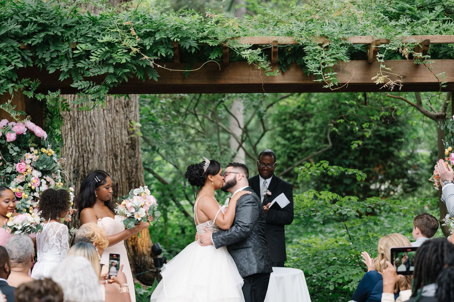 A couple shares their first kiss during an outdoor garden wedding ceremony surrounded by guests and lush greenery.
