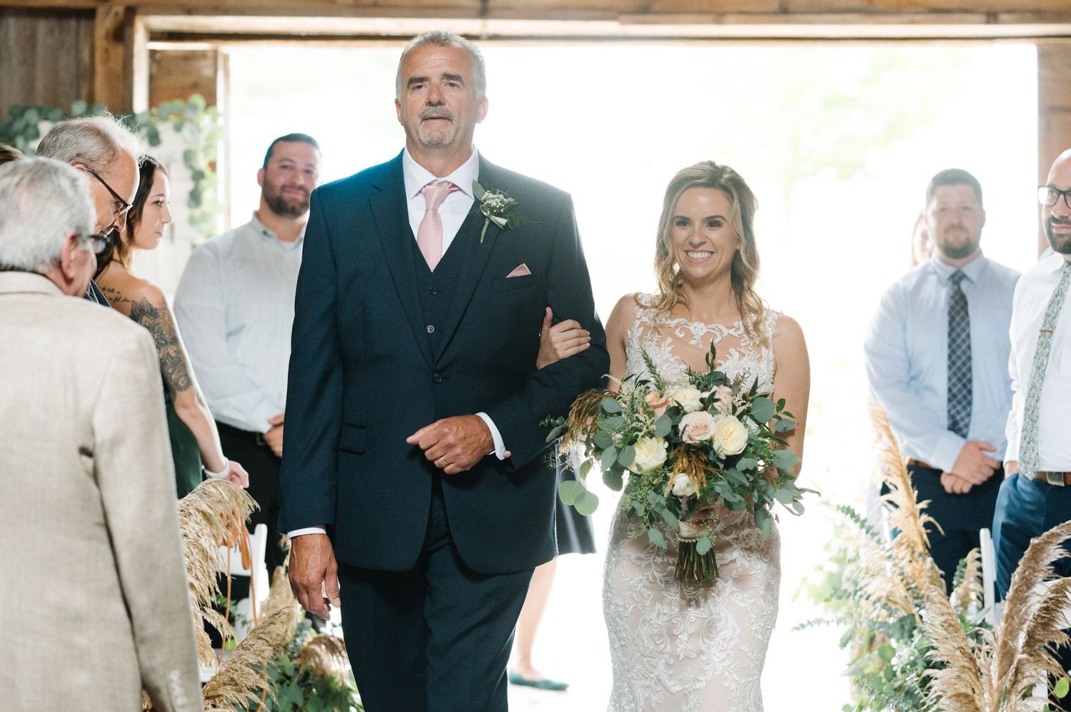A couple walks down the aisle at their outdoor wedding ceremony while carrying a bouquet of flowers and greenery.
