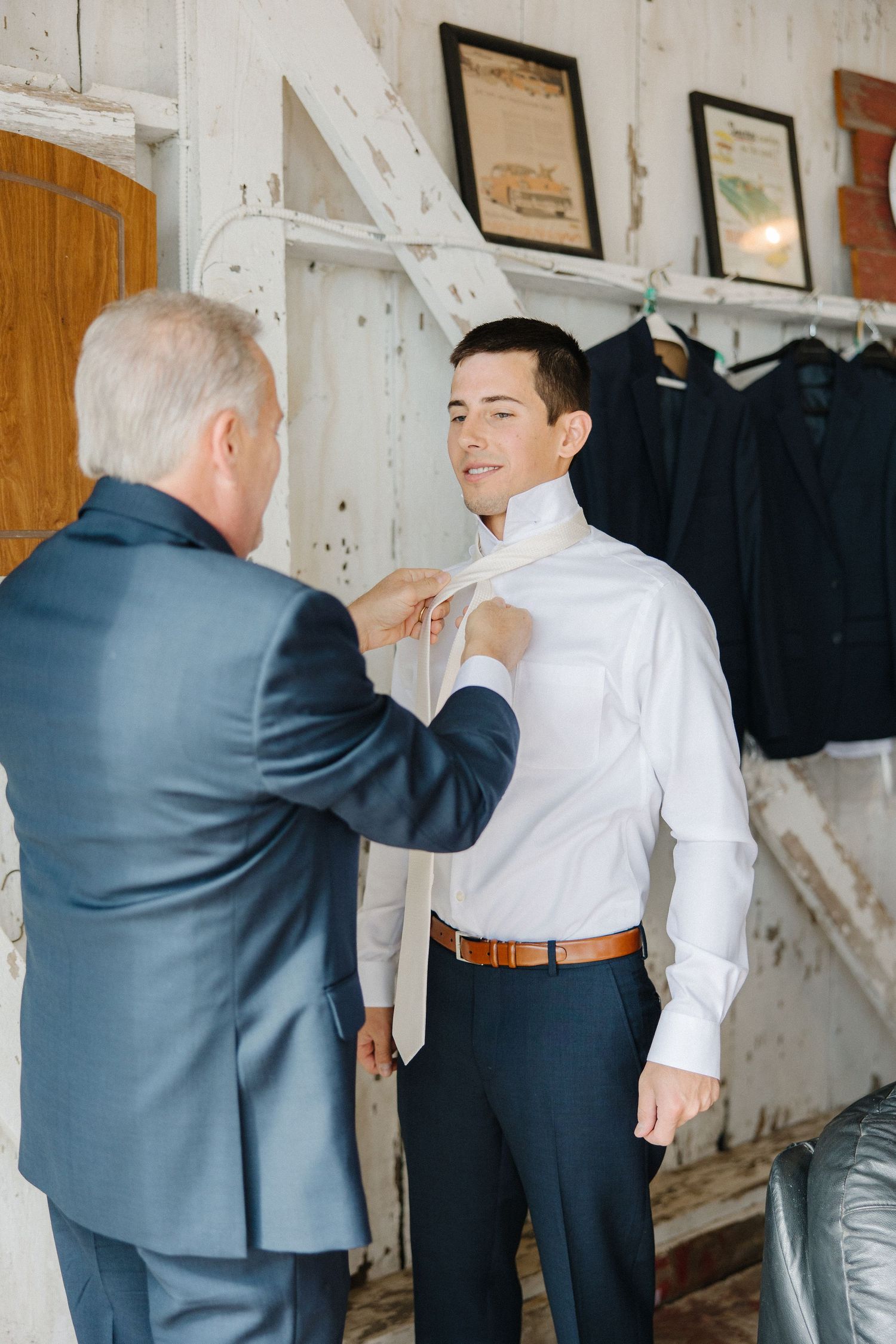 Two people in a dressing room as one helps adjust the other's formal attire and tie.