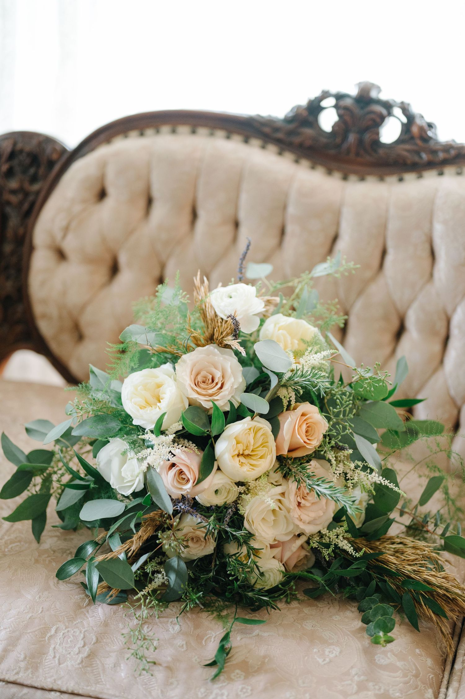A romantic floral bouquet with cream and blush roses nestled on a vintage tufted beige sofa chair.