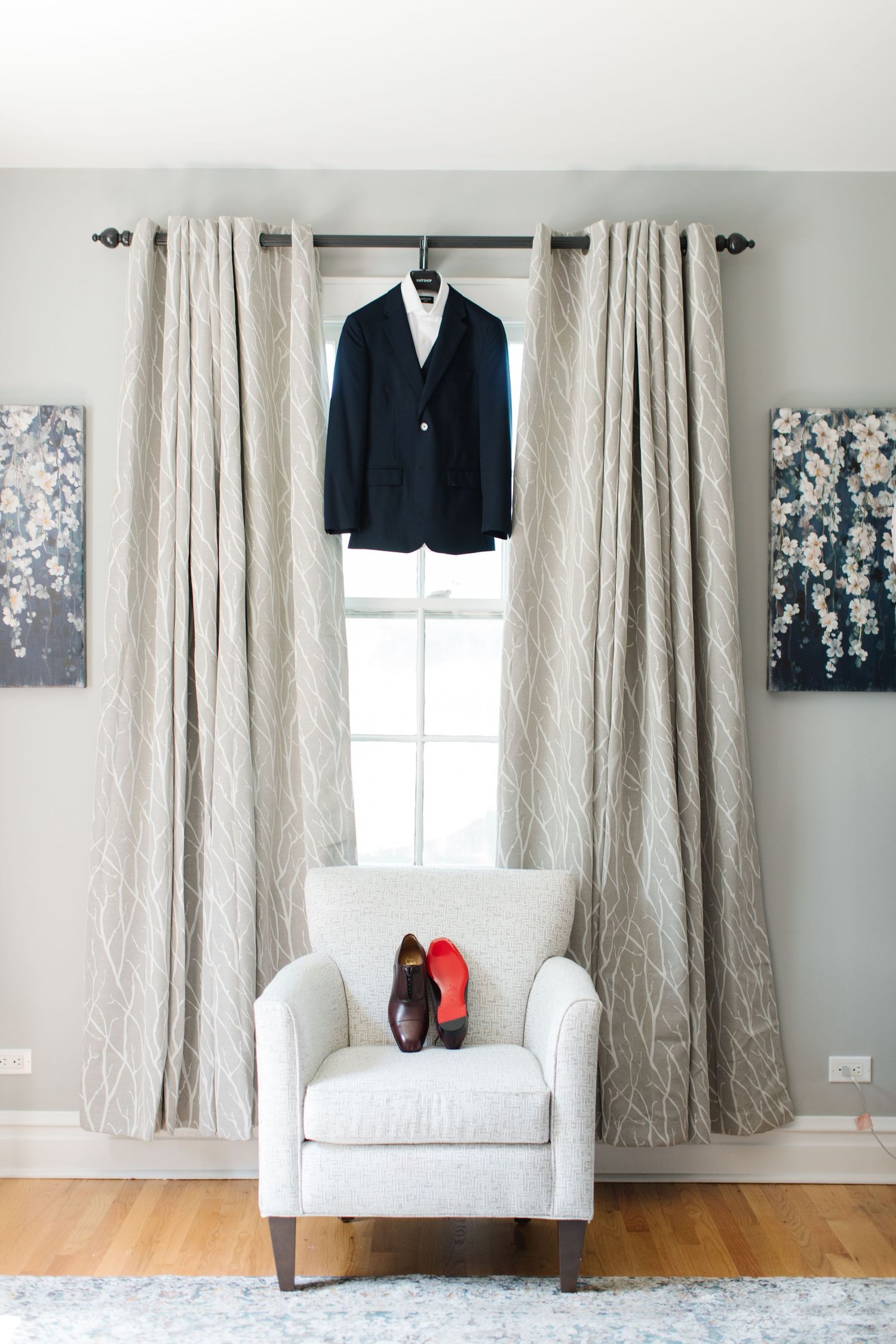 A navy blazer hangs above a white armchair with red shoes, framed by beige curtains and floral wall art.