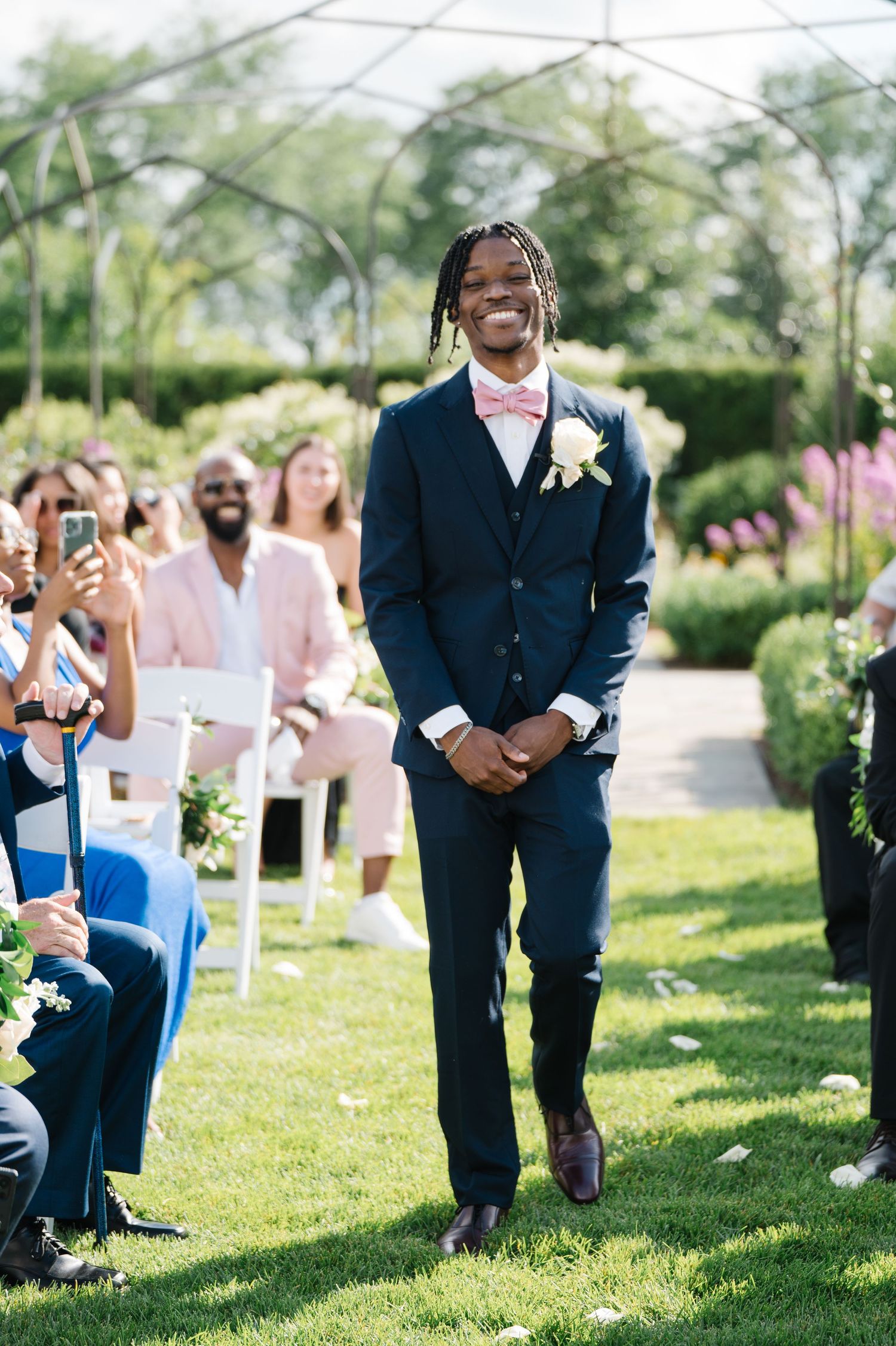 A groom stands at an outdoor garden wedding ceremony wearing a navy suit and boutonniere while guests look on.
