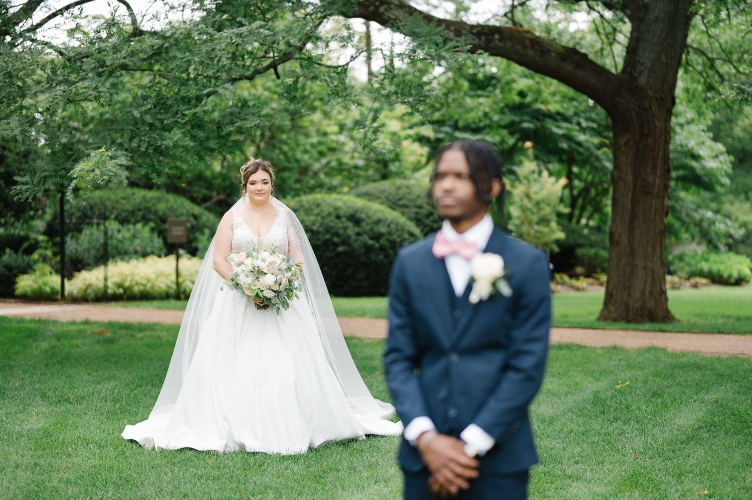 A bride and groom stand outdoors in a lush garden setting with large trees and green grass on their wedding day.