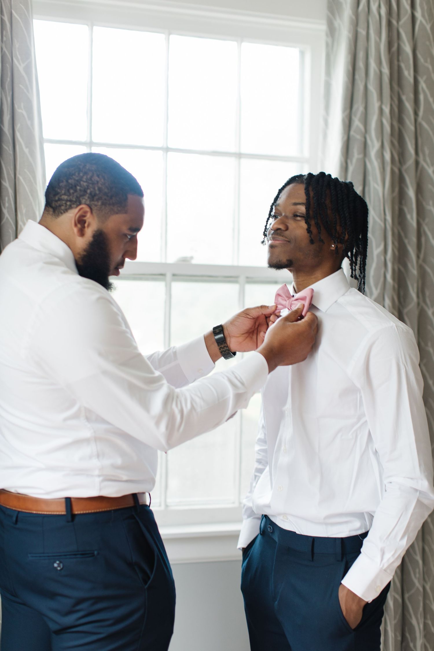 Two men in white shirts and navy pants get ready for a formal event as one adjusts the other's pink bow tie.