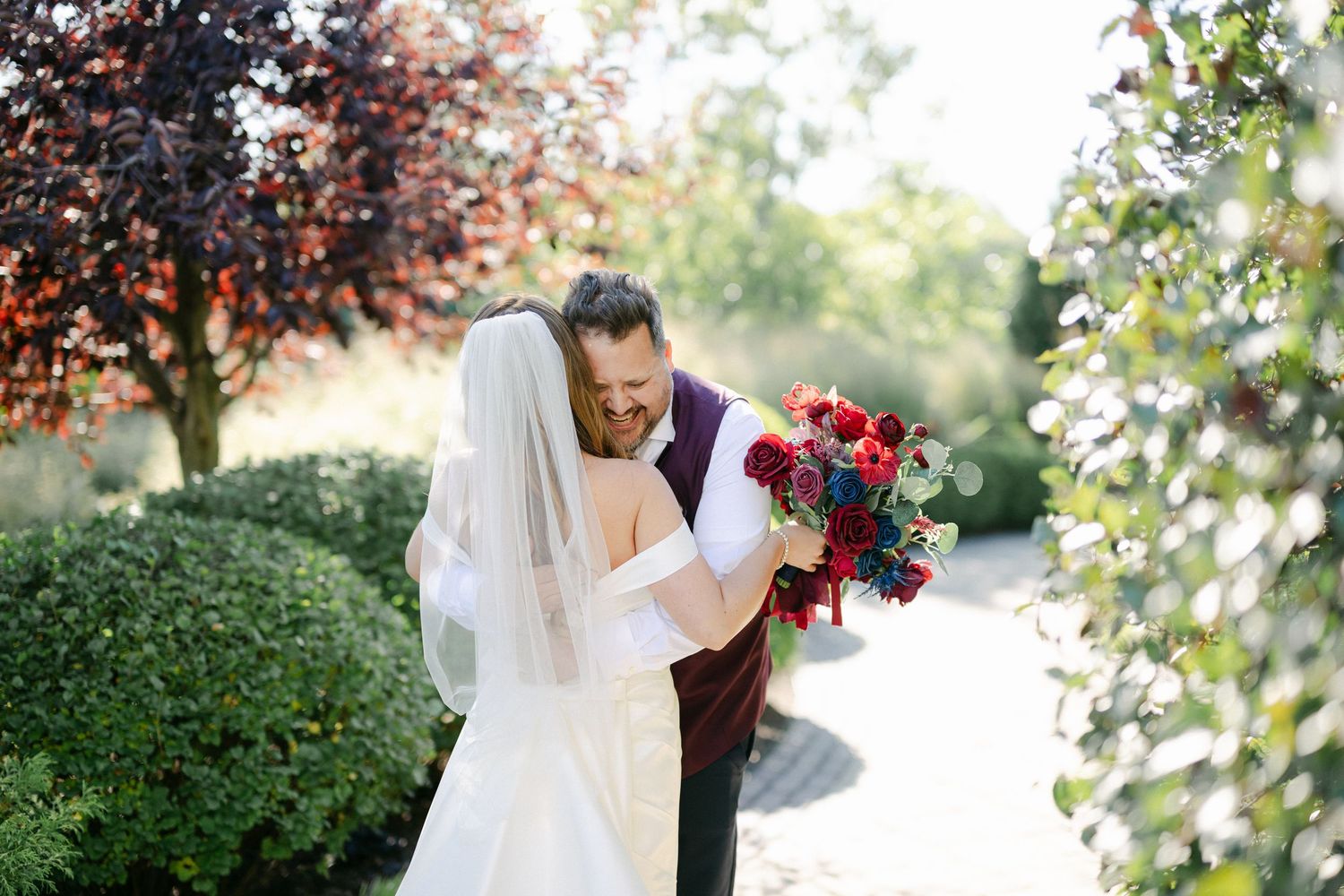 A bride in a white wedding dress holds a vibrant red and purple bouquet in a sunlit garden setting.