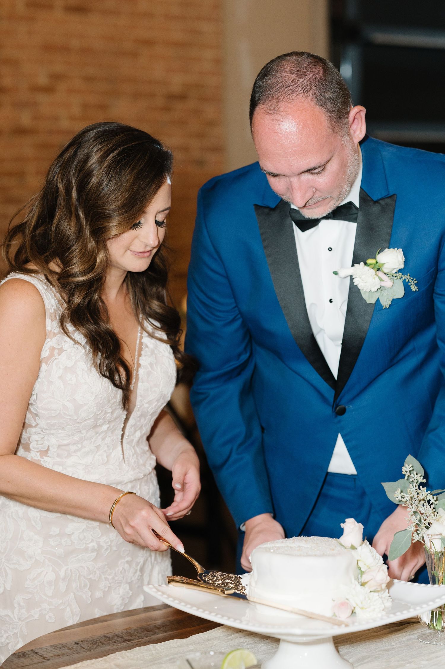 Couple in wedding attire cutting their wedding cake together, with bride in lace gown and groom in blue tuxedo jacket.