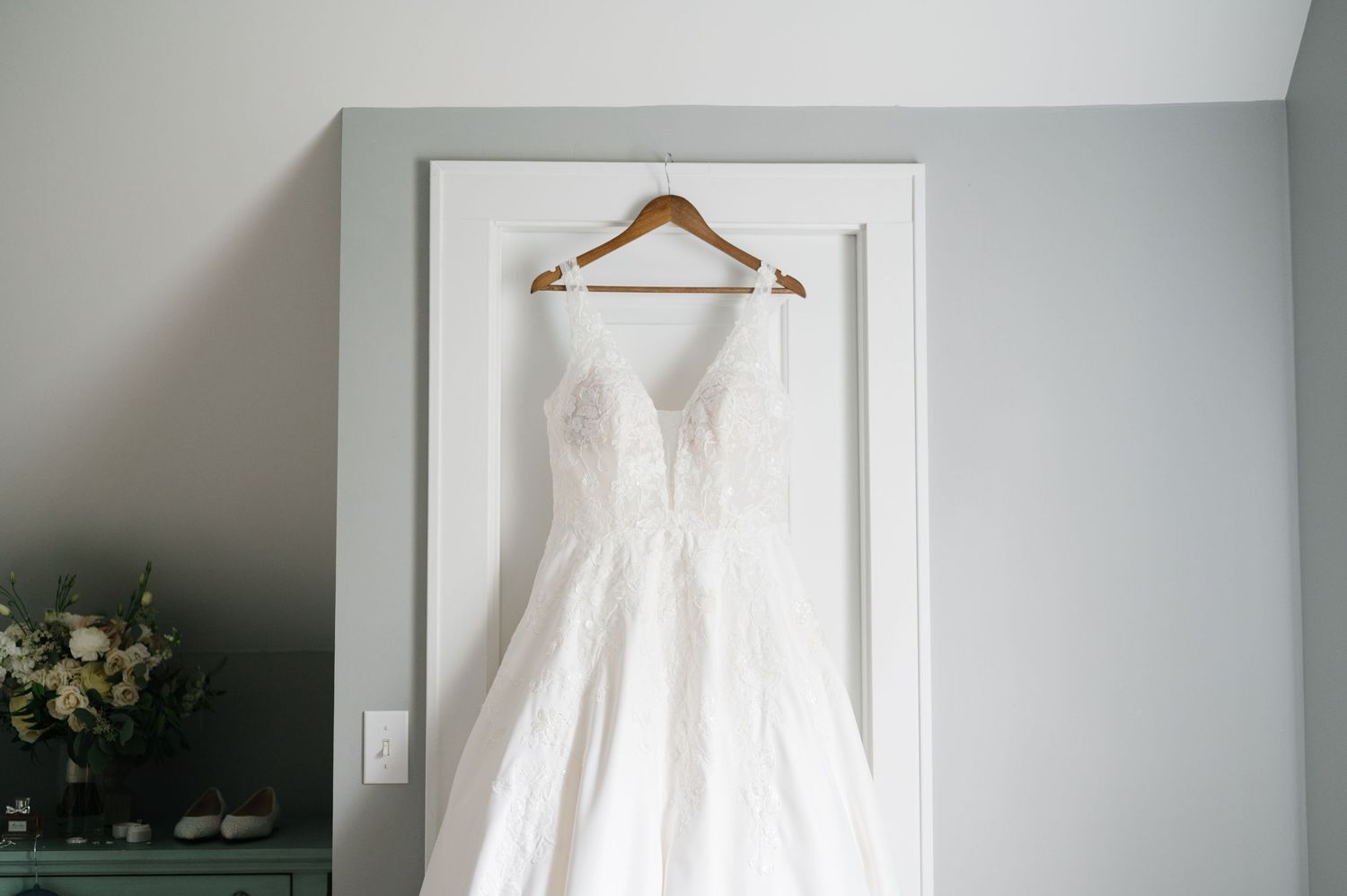 A white lace wedding dress hangs on a wooden hanger against a light gray wall, with flowers visible in the corner.