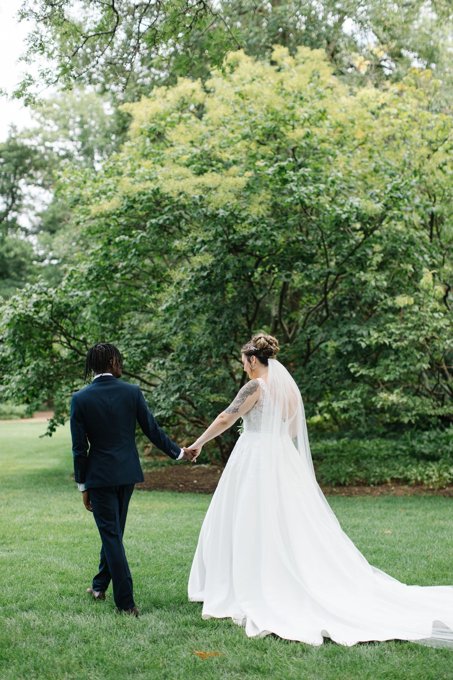 A couple in wedding attire walks hand in hand on a lush green lawn surrounded by trees on their special day.