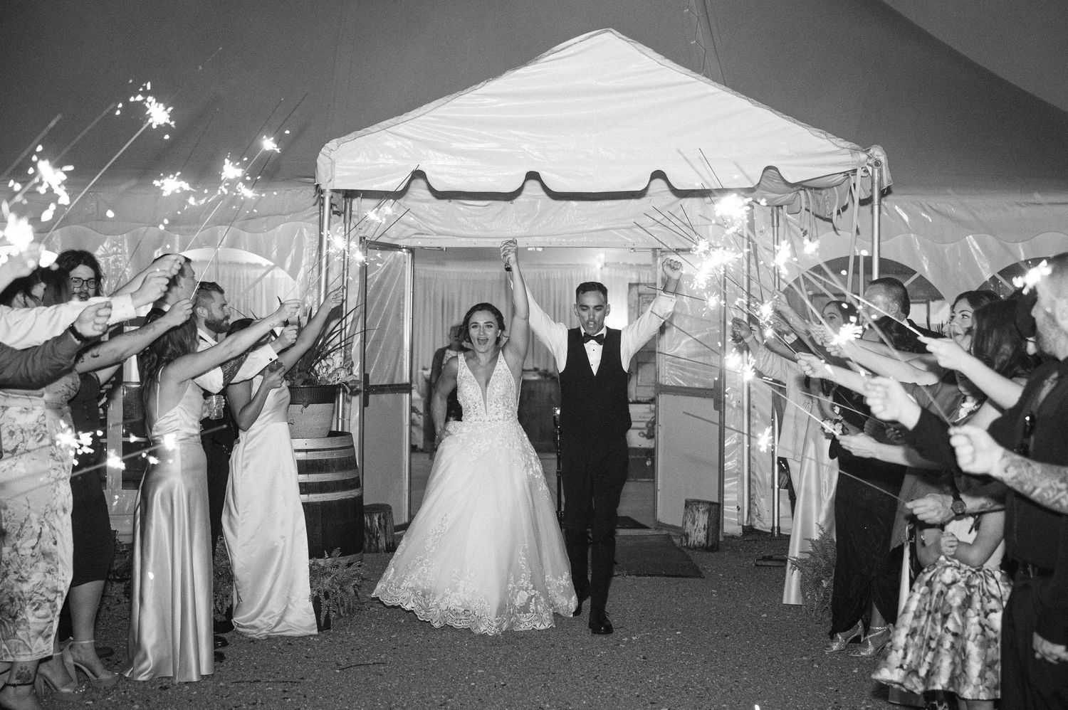 A joyful wedding exit under a white tent at night with sparklers lighting up the path for the newlyweds.