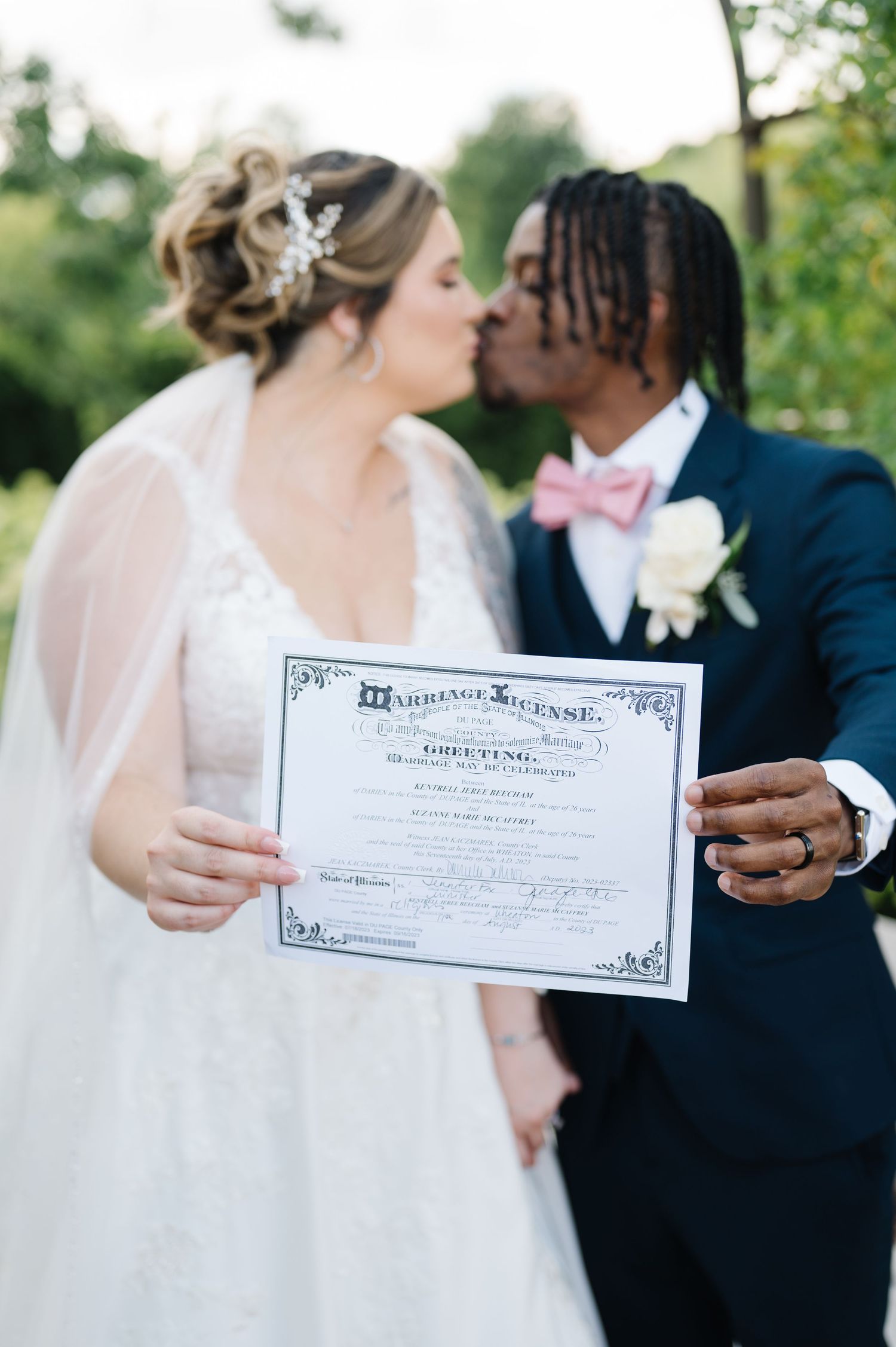 A bride and groom embrace while displaying their marriage certificate, with the groom wearing a pink bow tie and blue suit.