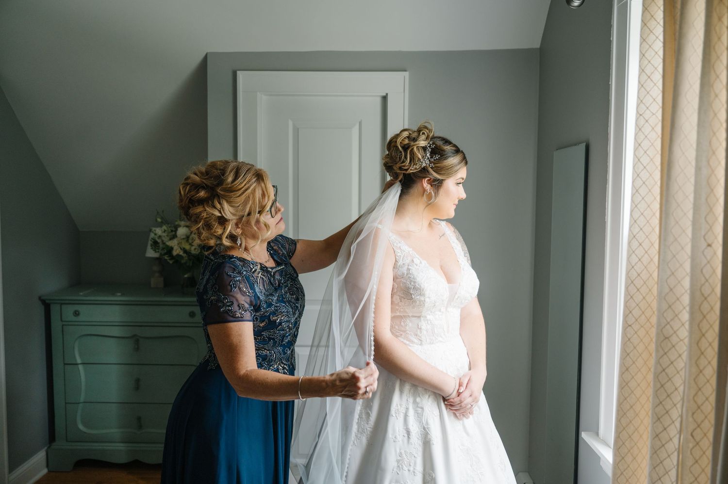 A bridal scene shows helping hands making final wedding dress adjustments in a softly lit bedroom.