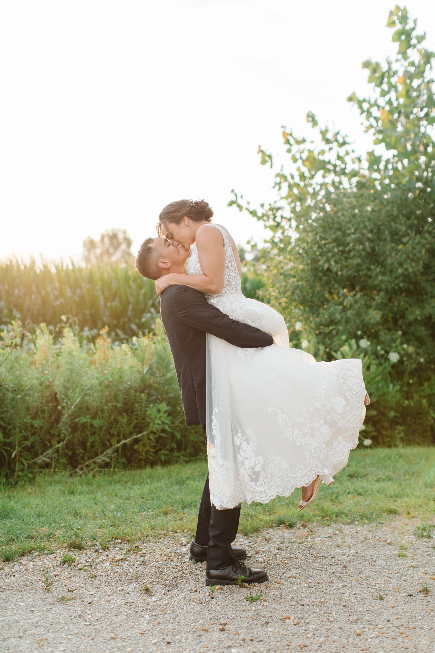 A romantic moment captured at sunset with gentle embrace and flowing white dress on a gravel path near greenery.