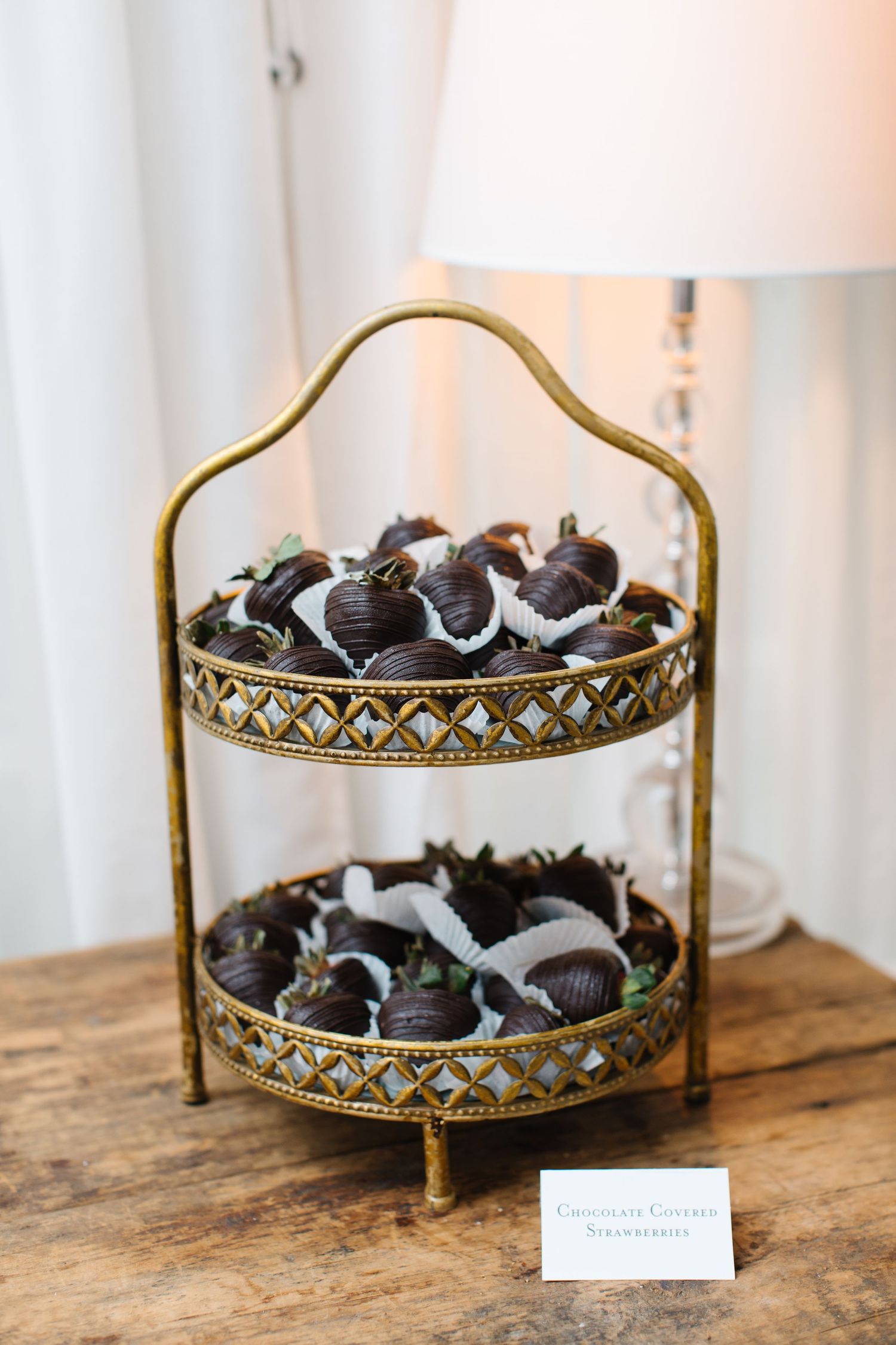 Chocolate covered Oreos displayed on an elegant two-tiered gold filigree stand on a wooden surface near a white lamp.