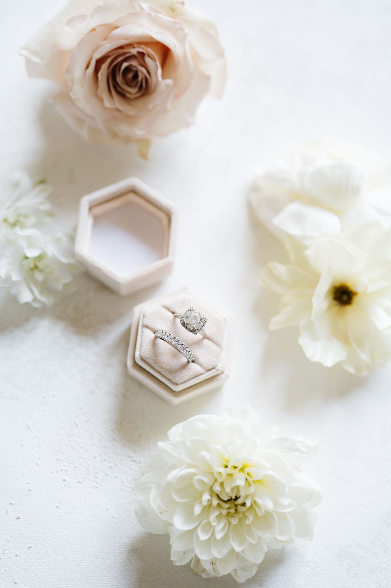 An elegant engagement ring in hexagonal box surrounded by soft white and blush flowers on white background.