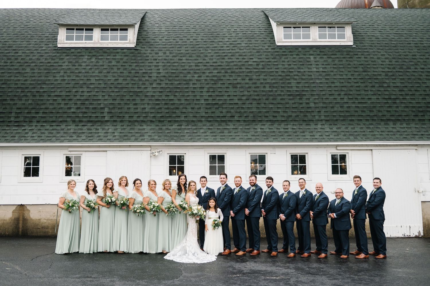 Wedding party poses in front of a white barn with green roof, bridesmaids in sage dresses and groomsmen in navy suits.