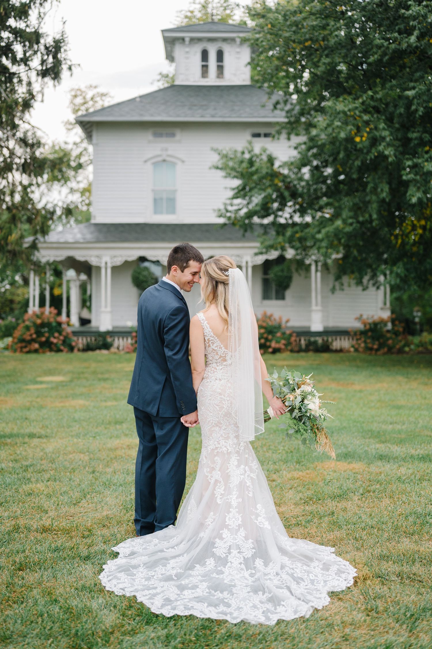 A romantic wedding photo of a couple in front of a beautiful white Victorian style mansion in full bloom.