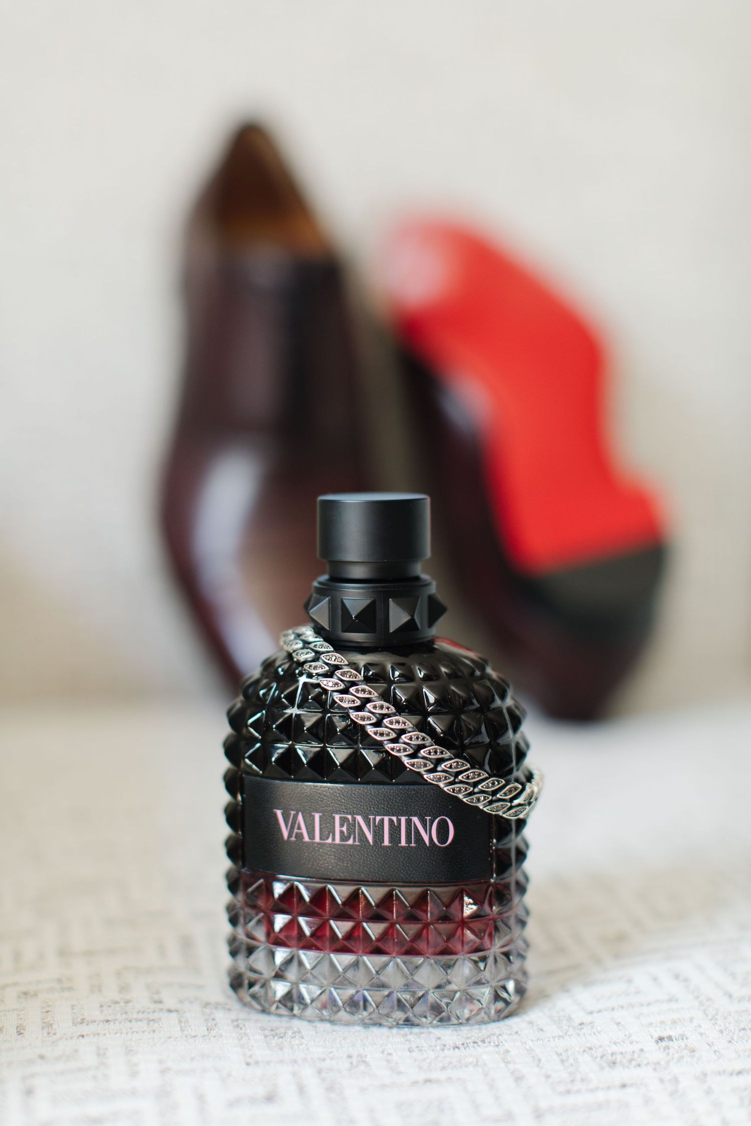 A dark Valentino perfume bottle with studded design and silver chain sits in front of a blurred shoe background.