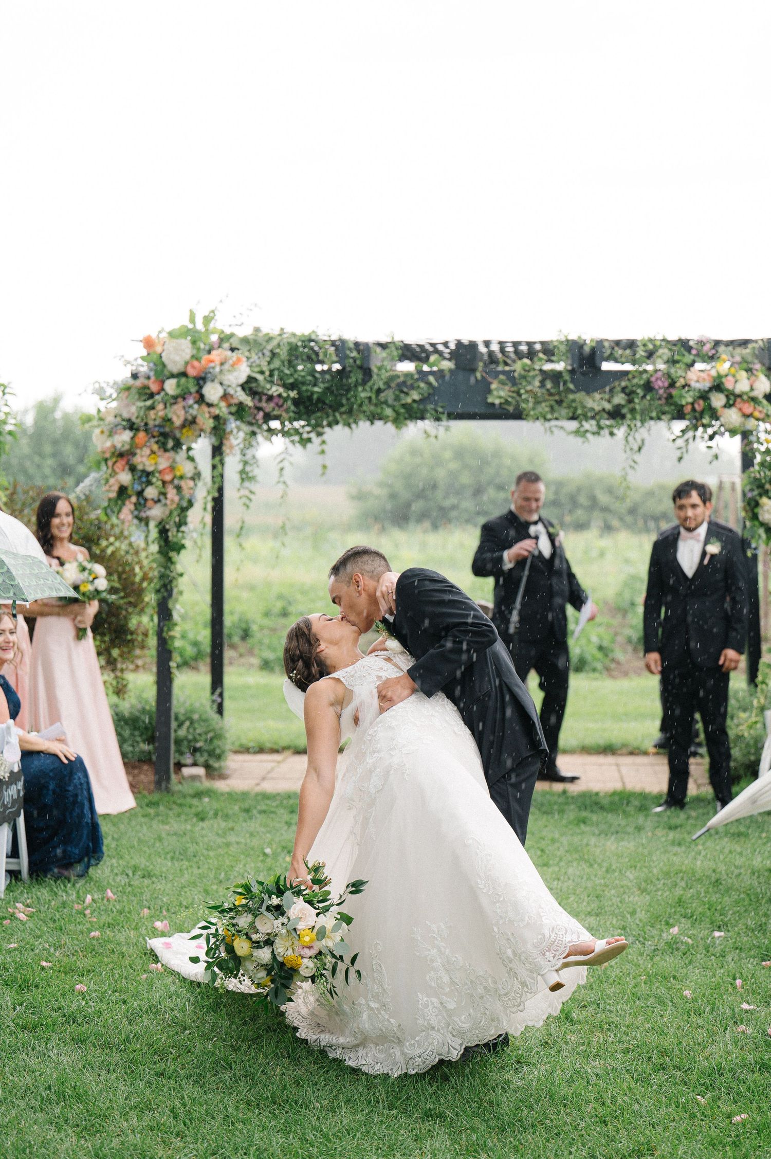 A romantic outdoor wedding ceremony features a couple embracing under a floral archway with guests looking on.