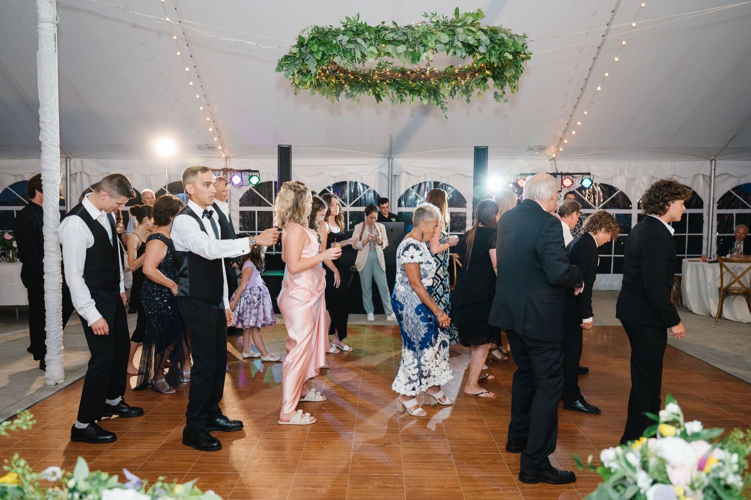 People dancing and socializing under a white tent with string lights and a green floral chandelier at an elegant event.