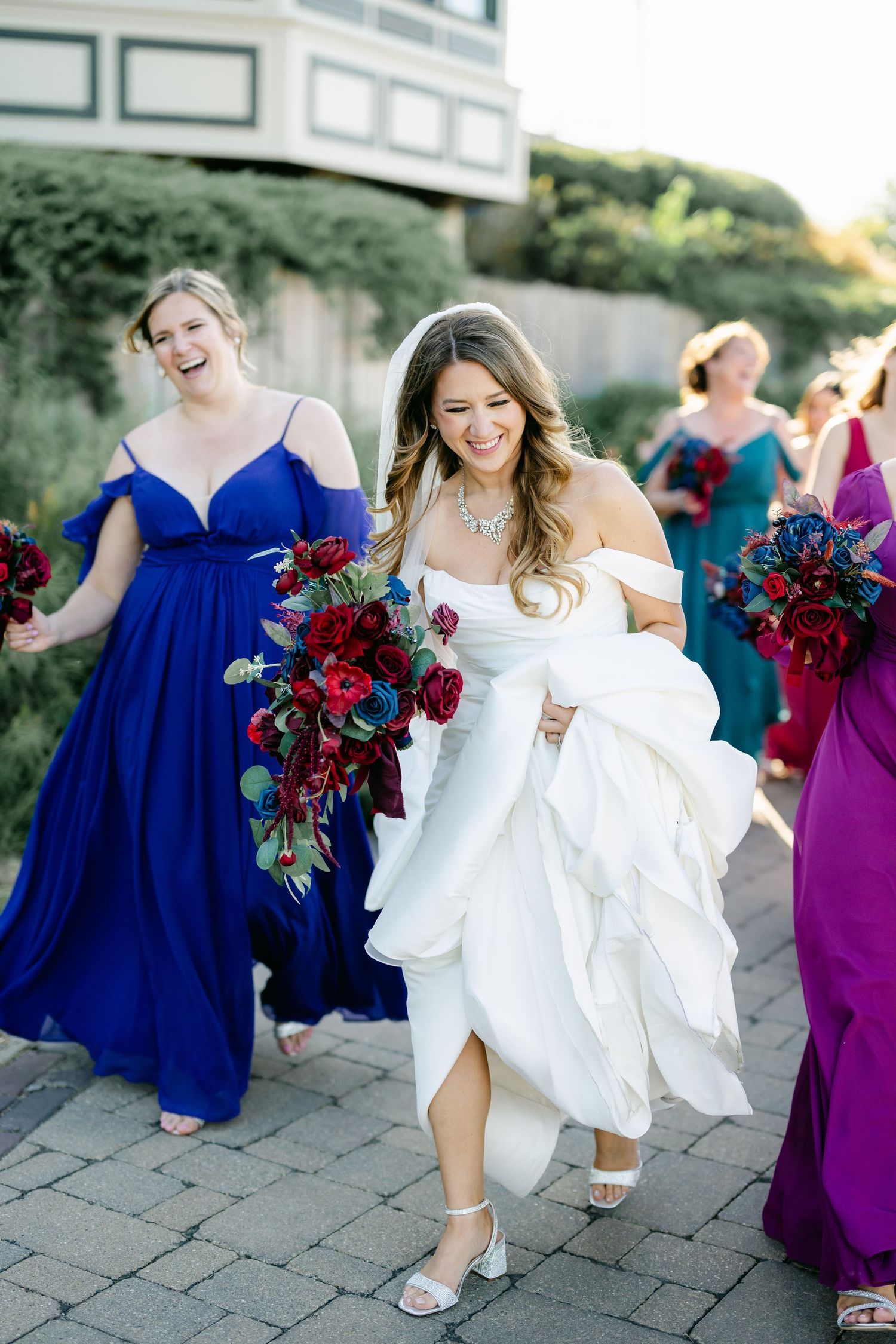 A bridal party walks outdoors with bridesmaids in jewel-toned dresses carrying rich burgundy and purple floral bouquets.