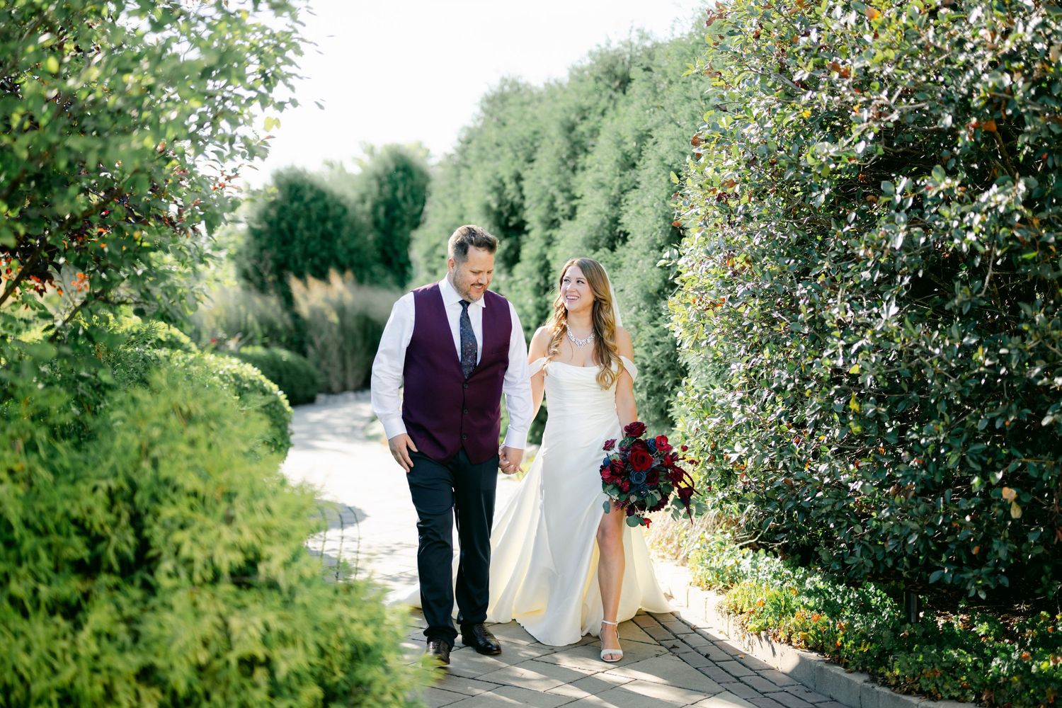 A couple in wedding attire walks along a winding garden path surrounded by lush greenery on a sunny day.