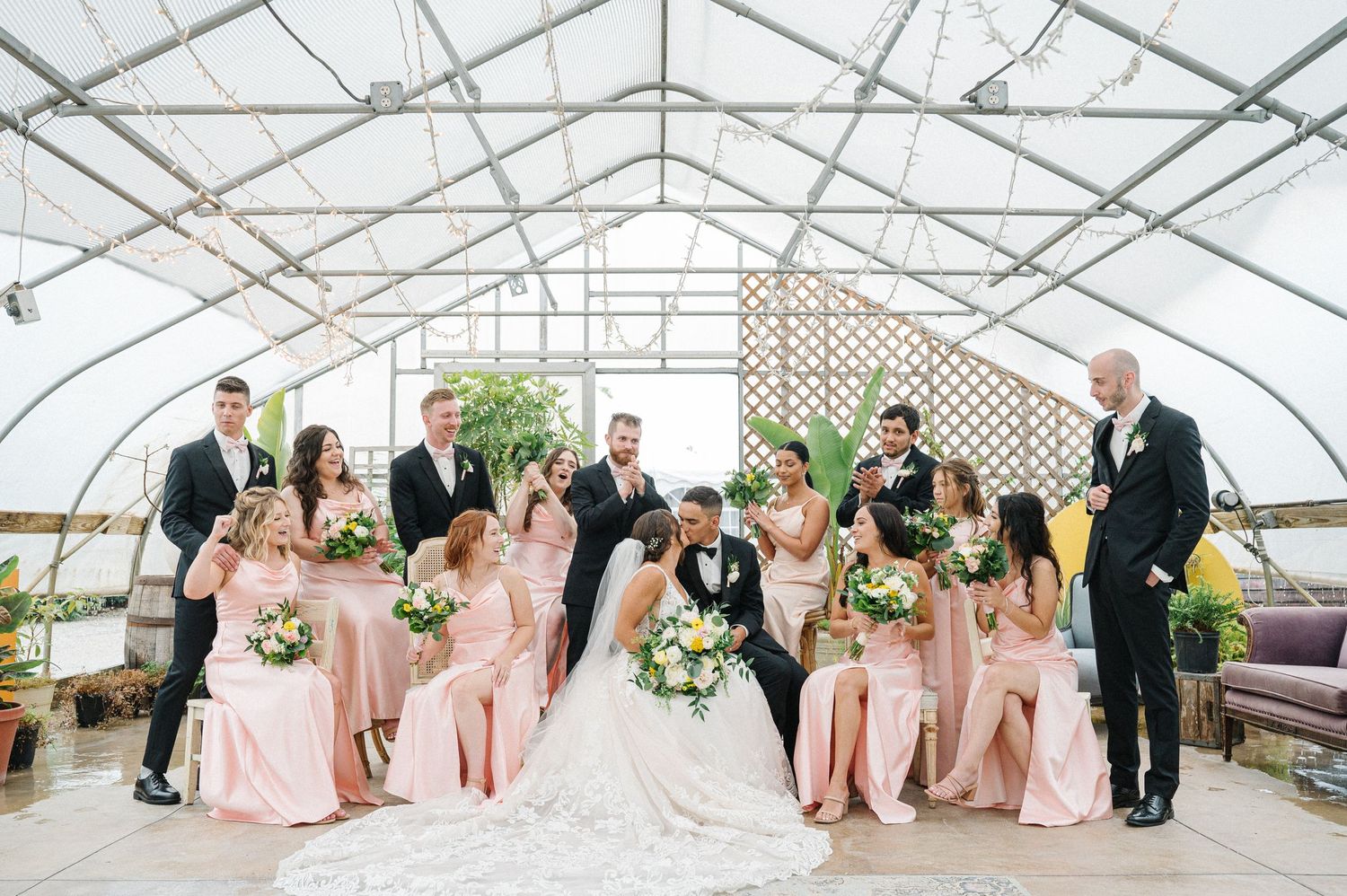 Wedding party poses together inside a bright greenhouse venue with pink bridesmaid dresses and formal attire.