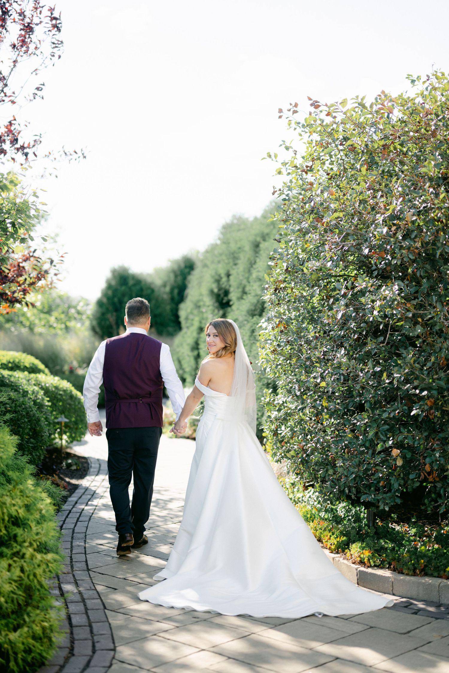 Newlyweds walk hand in hand down a winding garden path bordered by flowering bushes on a sunny afternoon.