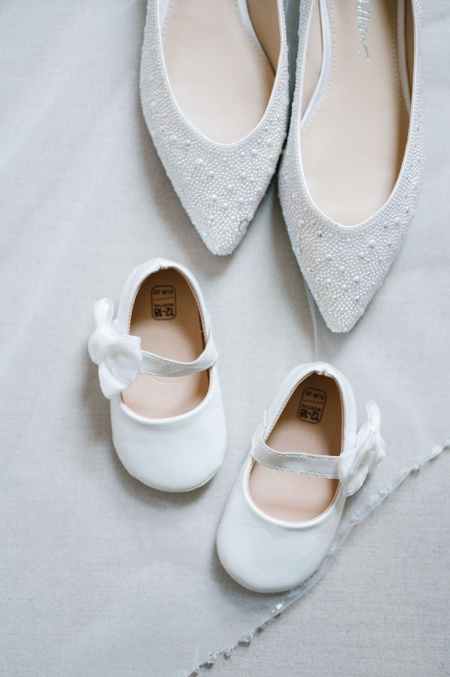 Elegant white bridal flats and beaded pointed-toe shoes arranged on light gray fabric background.