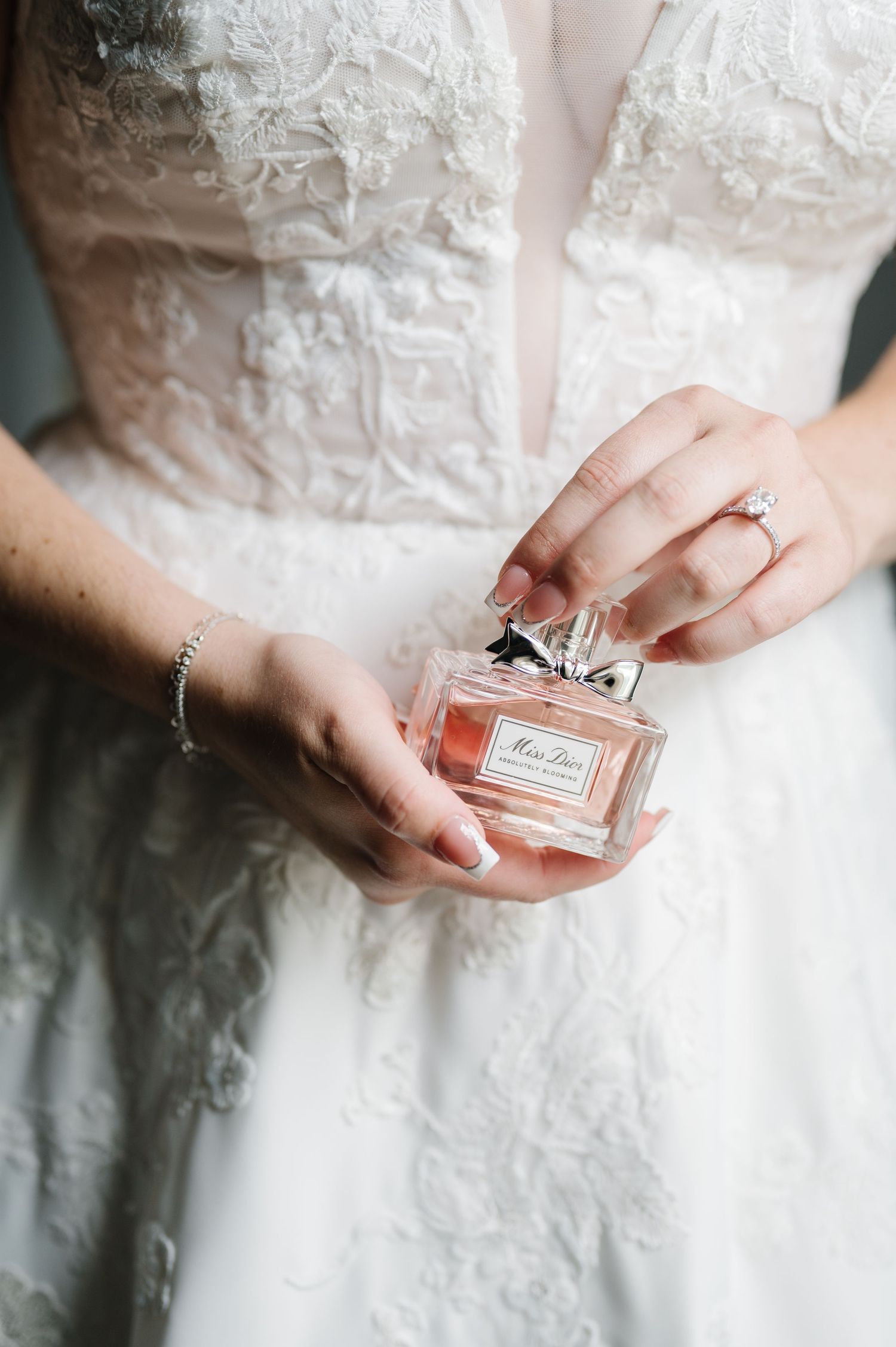 Close-up of hands holding Dior perfume bottle while wearing white lace wedding dress and engagement ring.