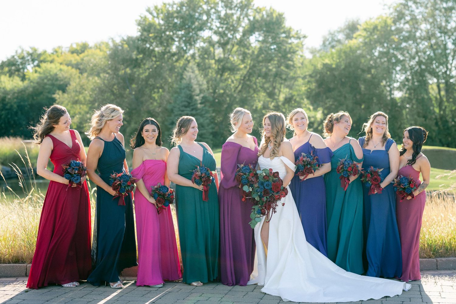 Wedding party stands outdoors wearing jewel-toned dresses in rich burgundy, emerald, and blue hues against a golden sunset.