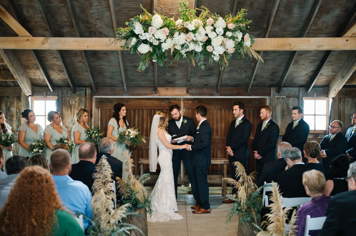 Rustic barn wedding ceremony with white floral arrangements hanging from wooden beams and guests seated on benches.