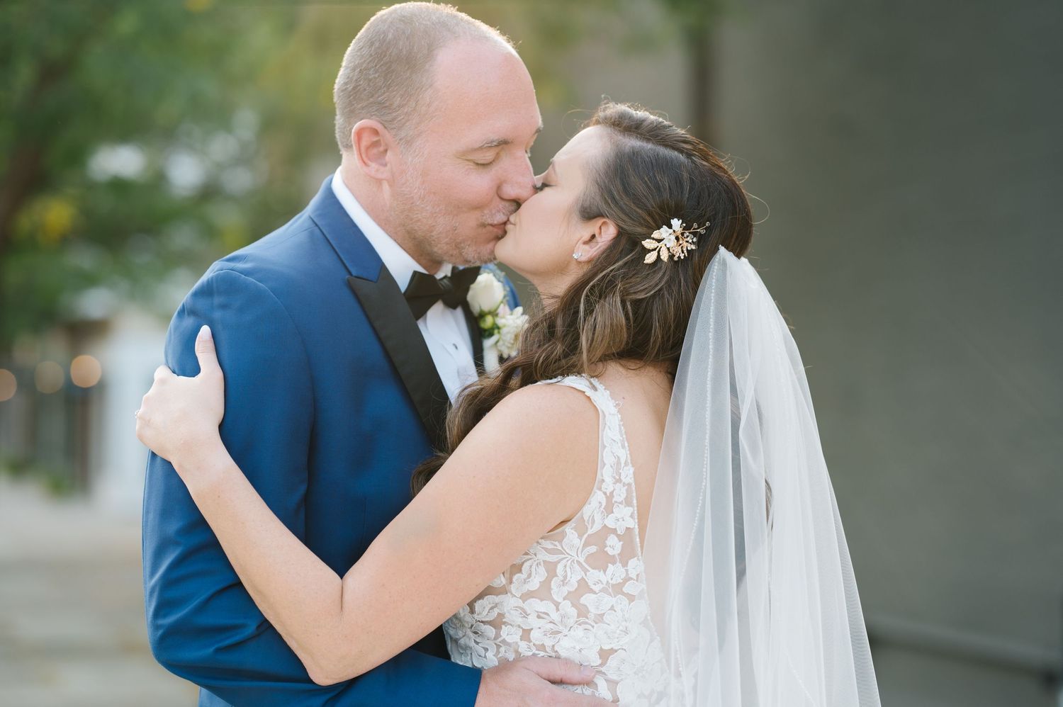 A romantic wedding couple in a navy suit and lace gown share a tender kiss outdoors.