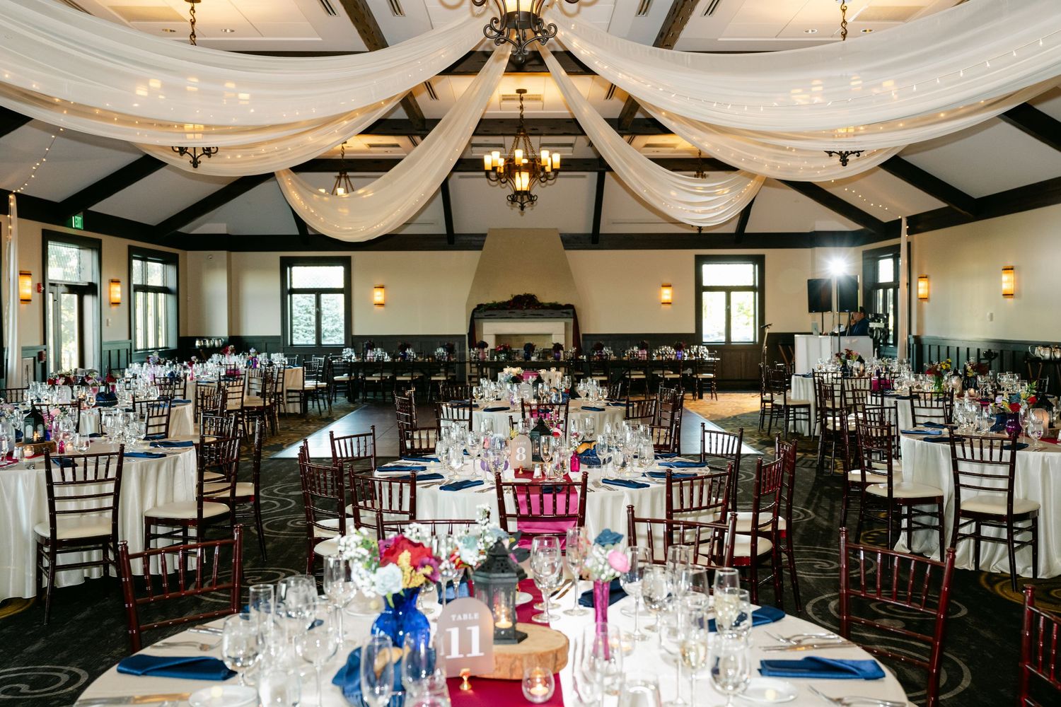 Elegant wedding reception venue with draped fabric ceiling, dark wood chiavari chairs, and colorful floral centerpieces.