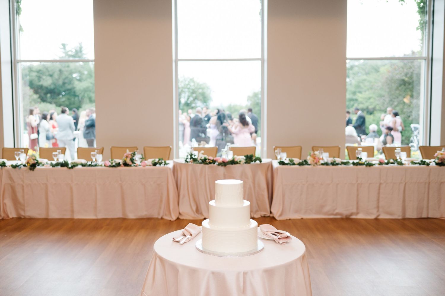 Elegant white wedding cake on round table with long banquet tables decorated in white linens behind large windows.