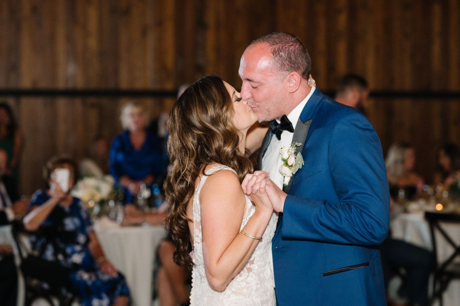 A couple shares their first dance together at a wedding reception in a rustic barn venue with guests in the background.