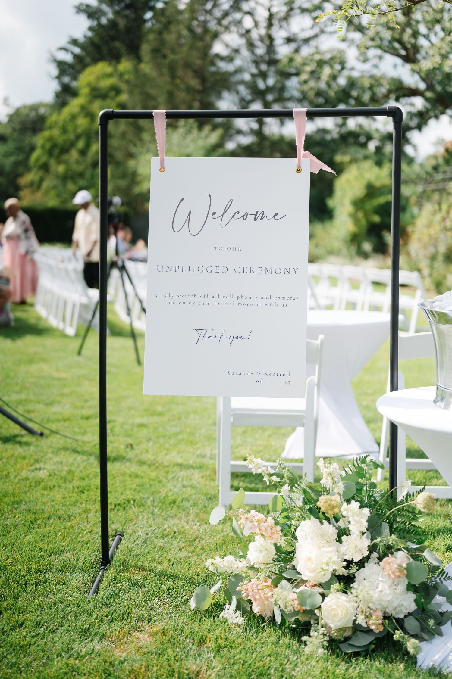 A welcome sign for an outdoor ceremony hangs on a black frame with pink clips, adorned by floral arrangements on the lawn.