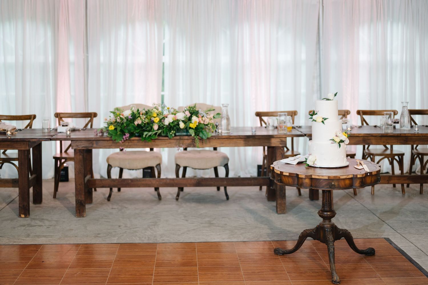 A rustic reception setup with wooden farm tables, cross-back chairs, and an elegant white wedding cake on a pedestal table.