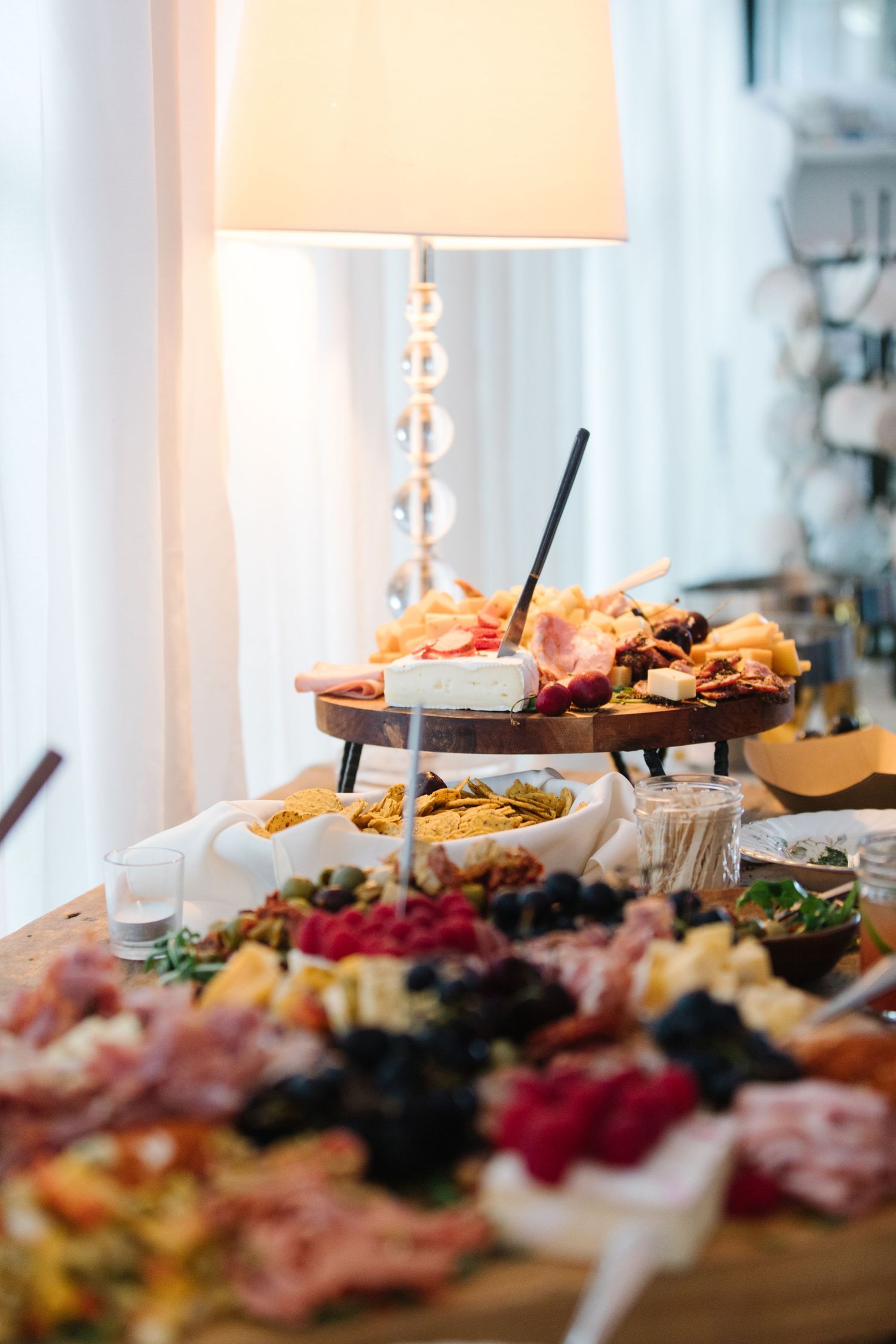 Elegant charcuterie and appetizer spread on wooden table with glass lamp and soft ambient lighting.