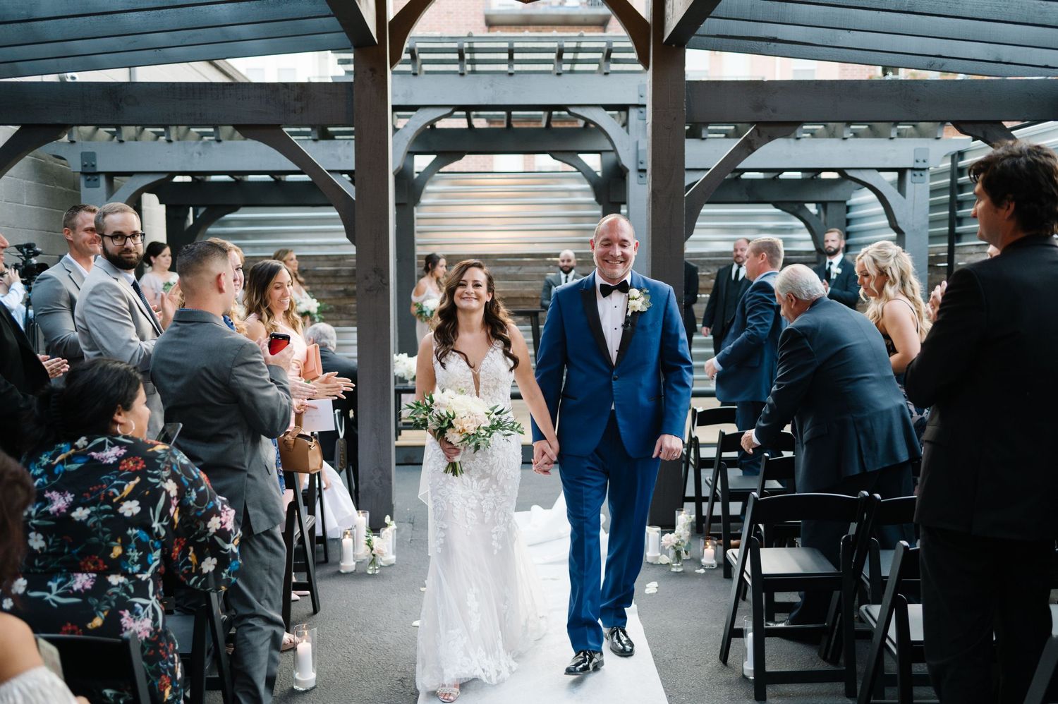 A wedding ceremony takes place under a covered outdoor venue with guests seated on black chairs.