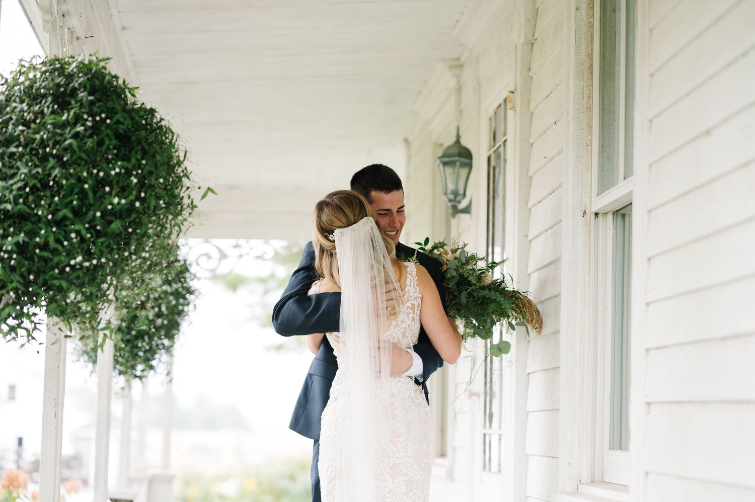 A couple embraces on a white columned porch with hanging plants and vintage lantern during their wedding day.