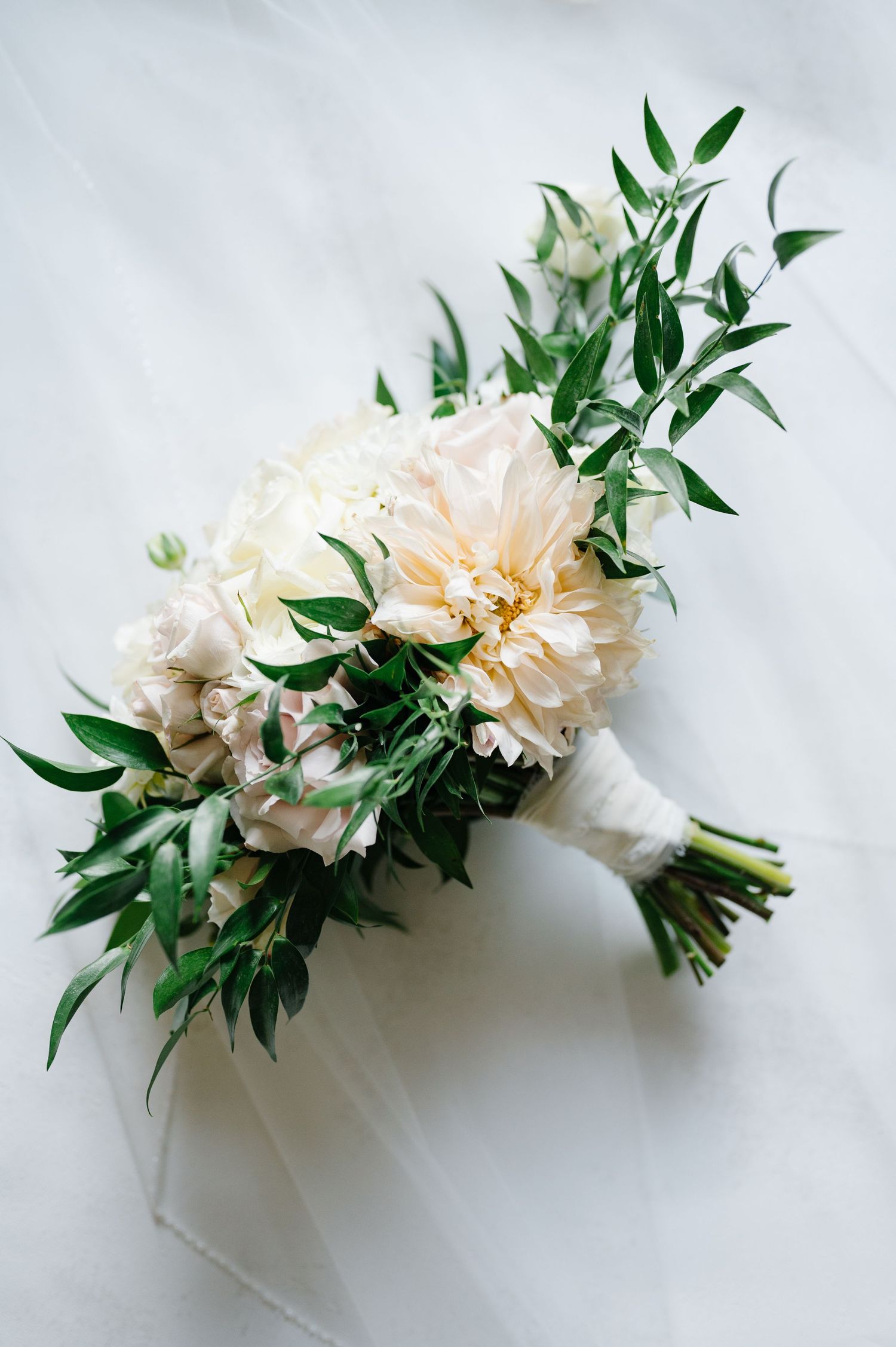A delicate bridal bouquet featuring soft peach peonies and fresh green olive leaves tied with white ribbon.