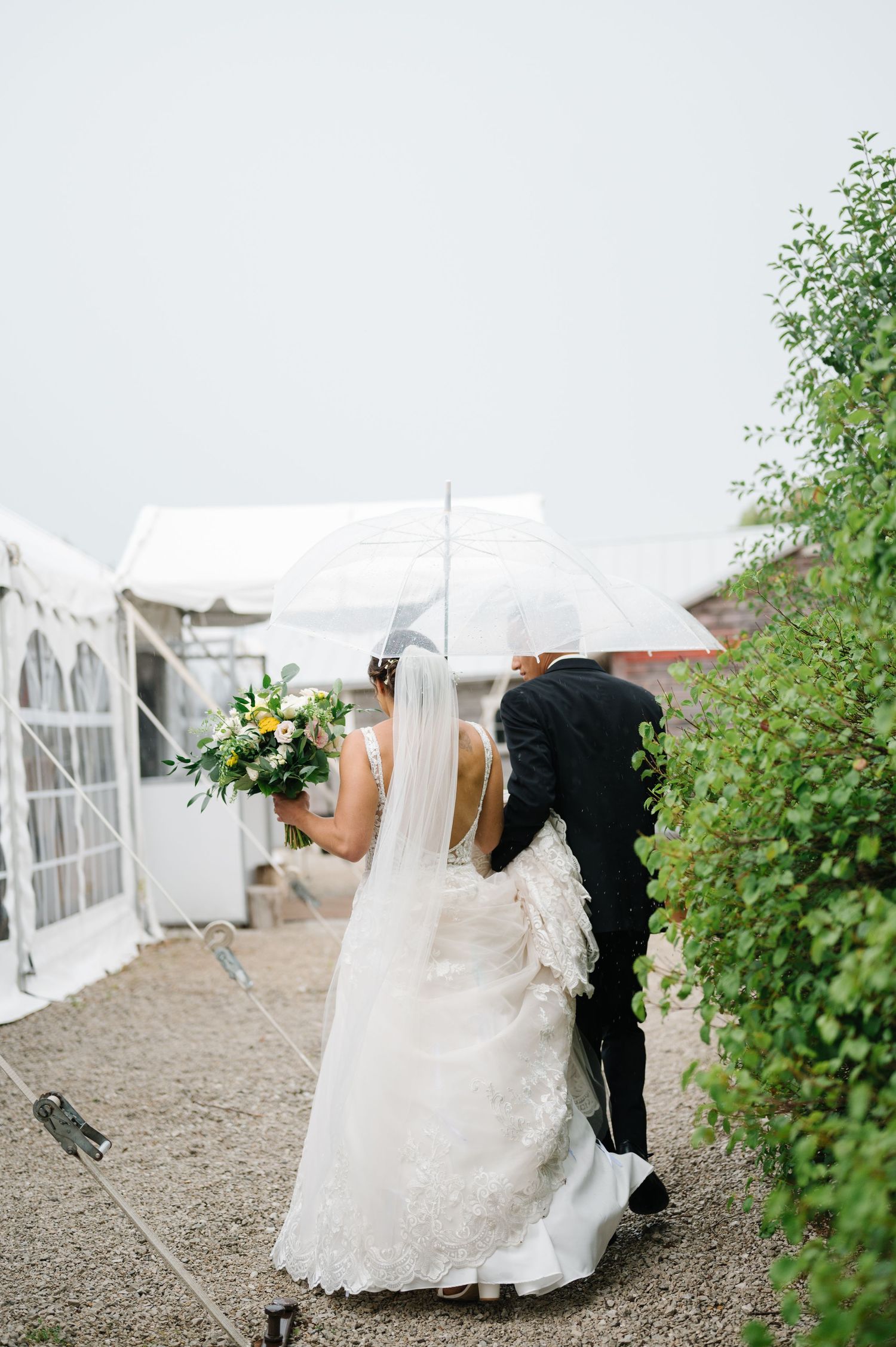A bride in a white lace dress walks with someone holding flowers beside a white tent at an outdoor wedding celebration.