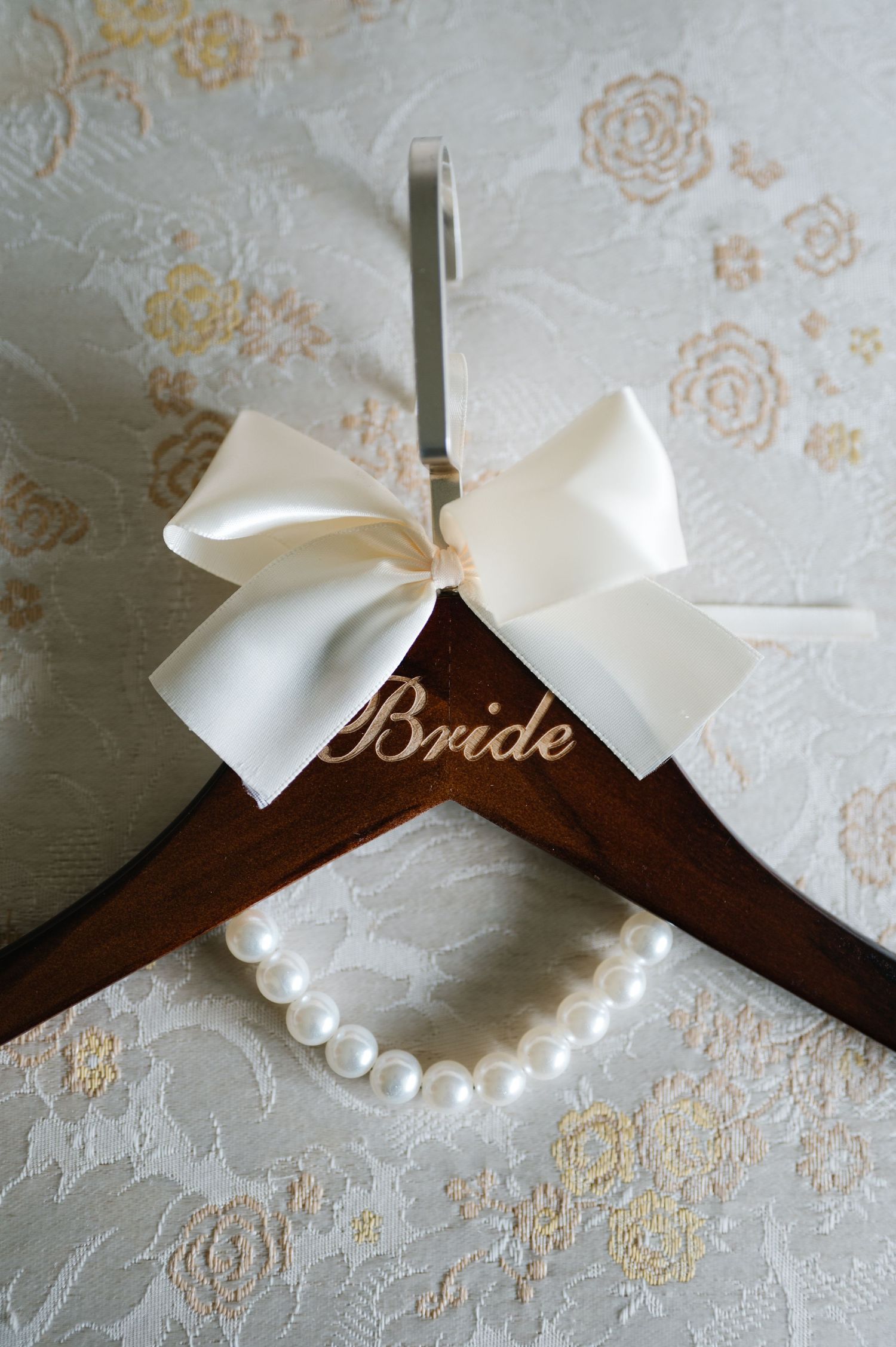 Wooden bridal hanger decorated with a cream satin bow and pearl necklace on floral lace background.