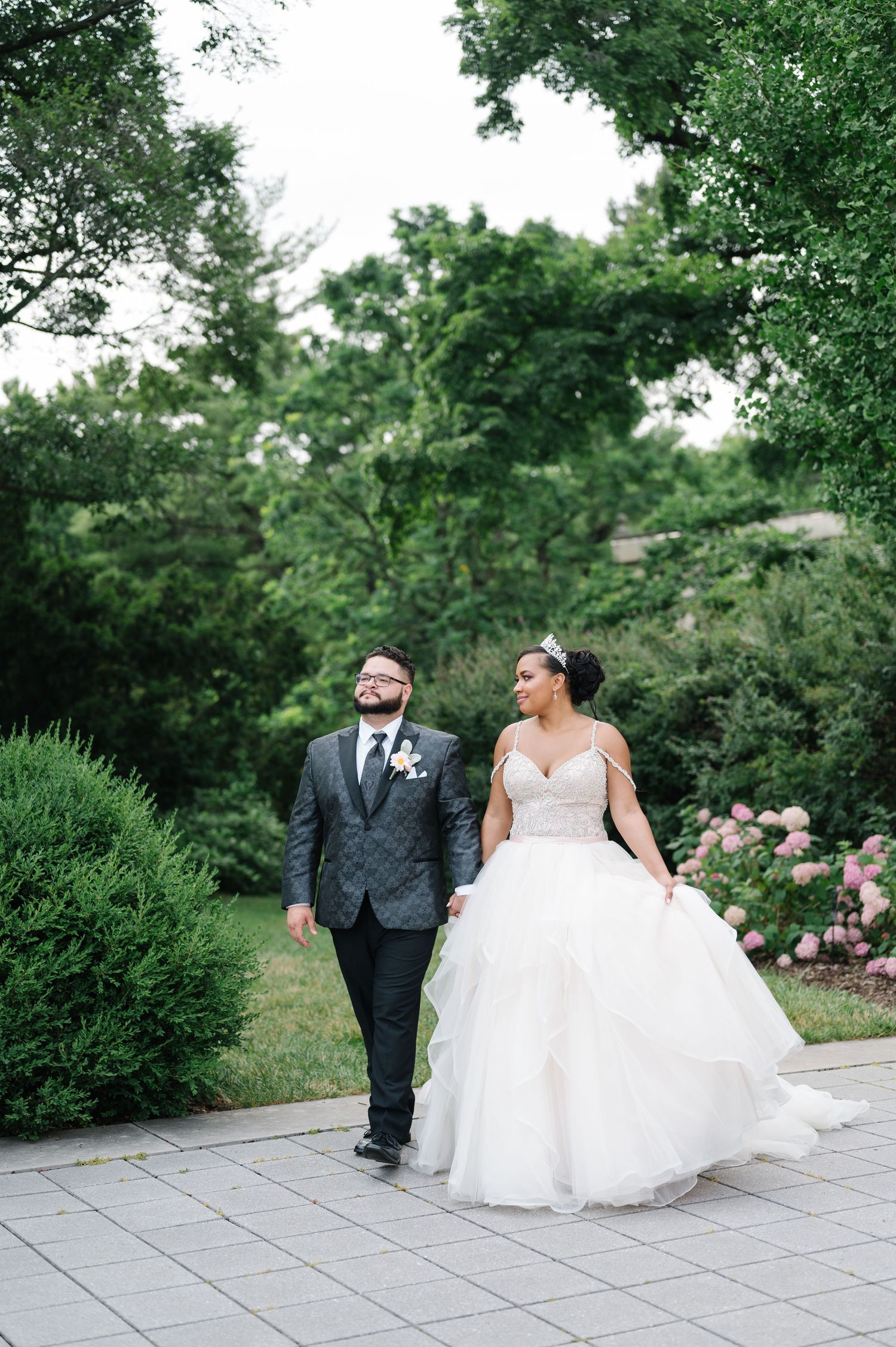 A couple walking on a stone patio surrounded by lush gardens with pink flowers and greenery on their wedding day.