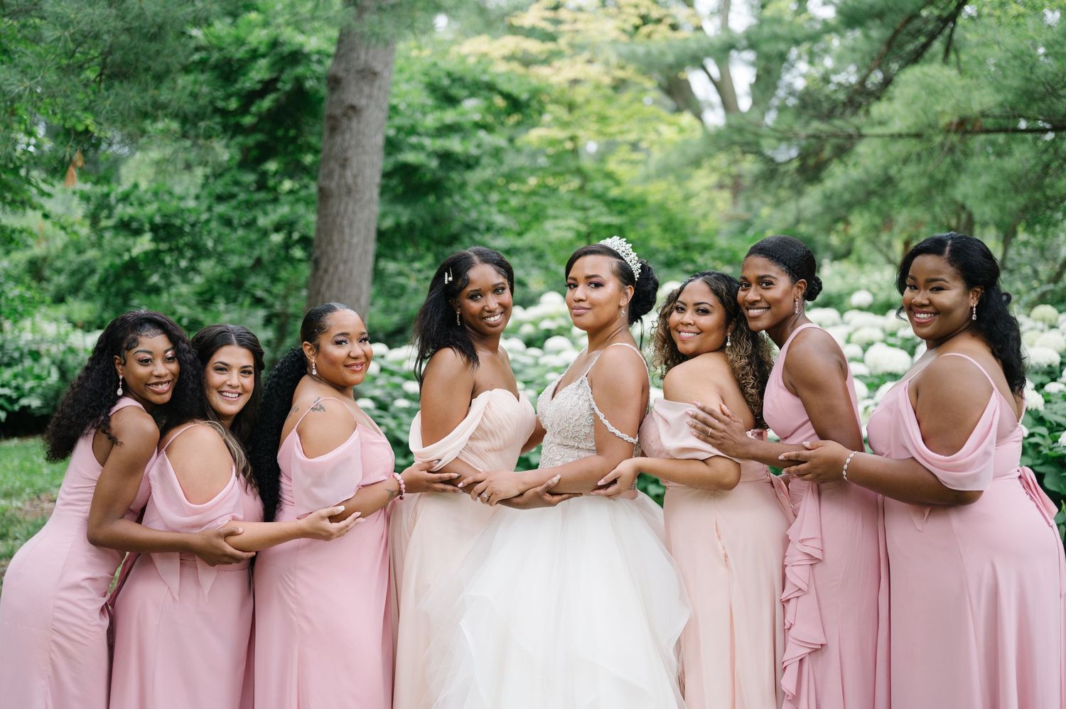 A wedding party wearing pink bridesmaid dresses and a white bridal gown stand close together in a lush garden setting.
