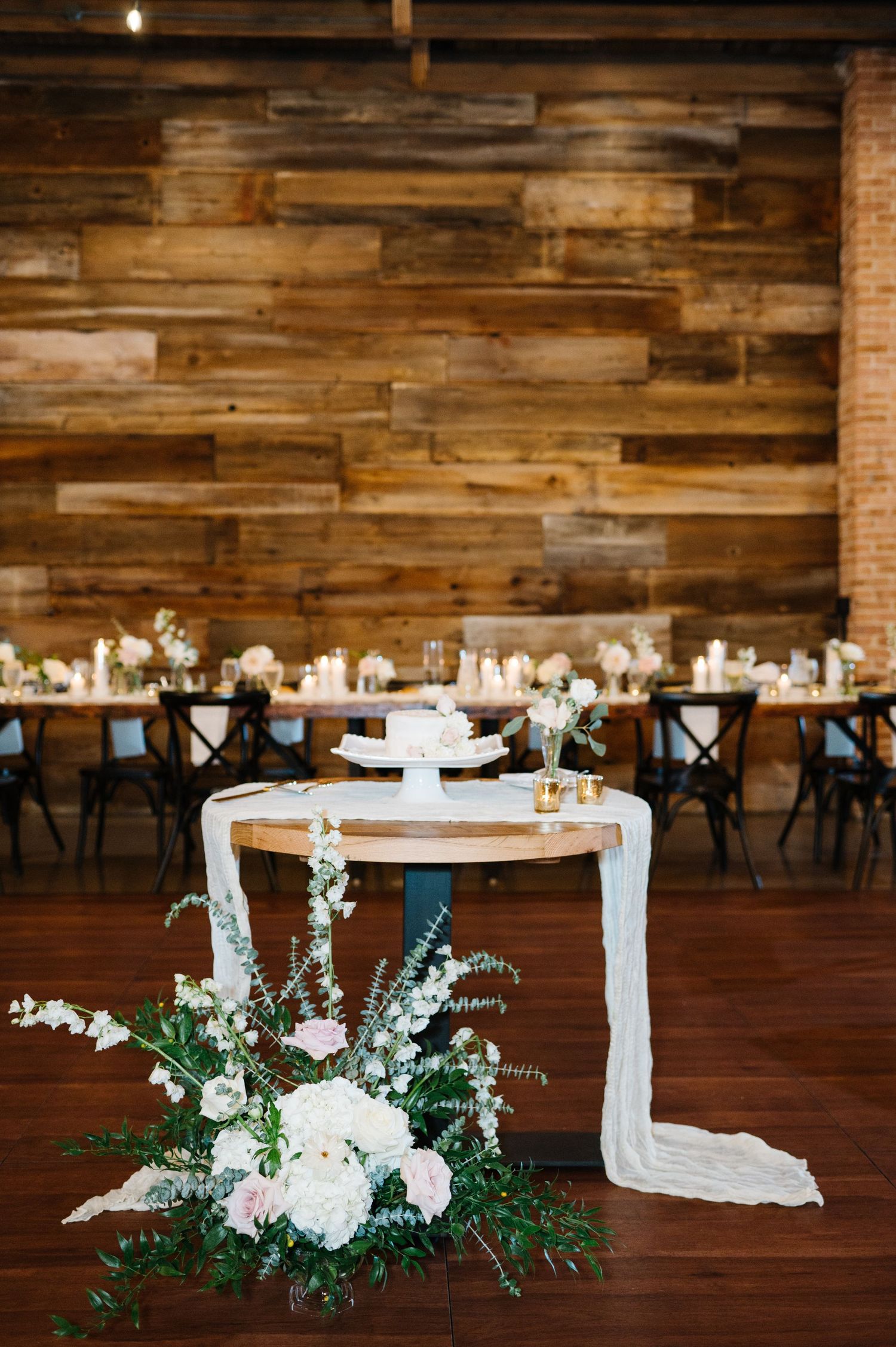 Rustic wedding reception setup with white floral arrangement, draped cake table and wood panel wall backdrop.