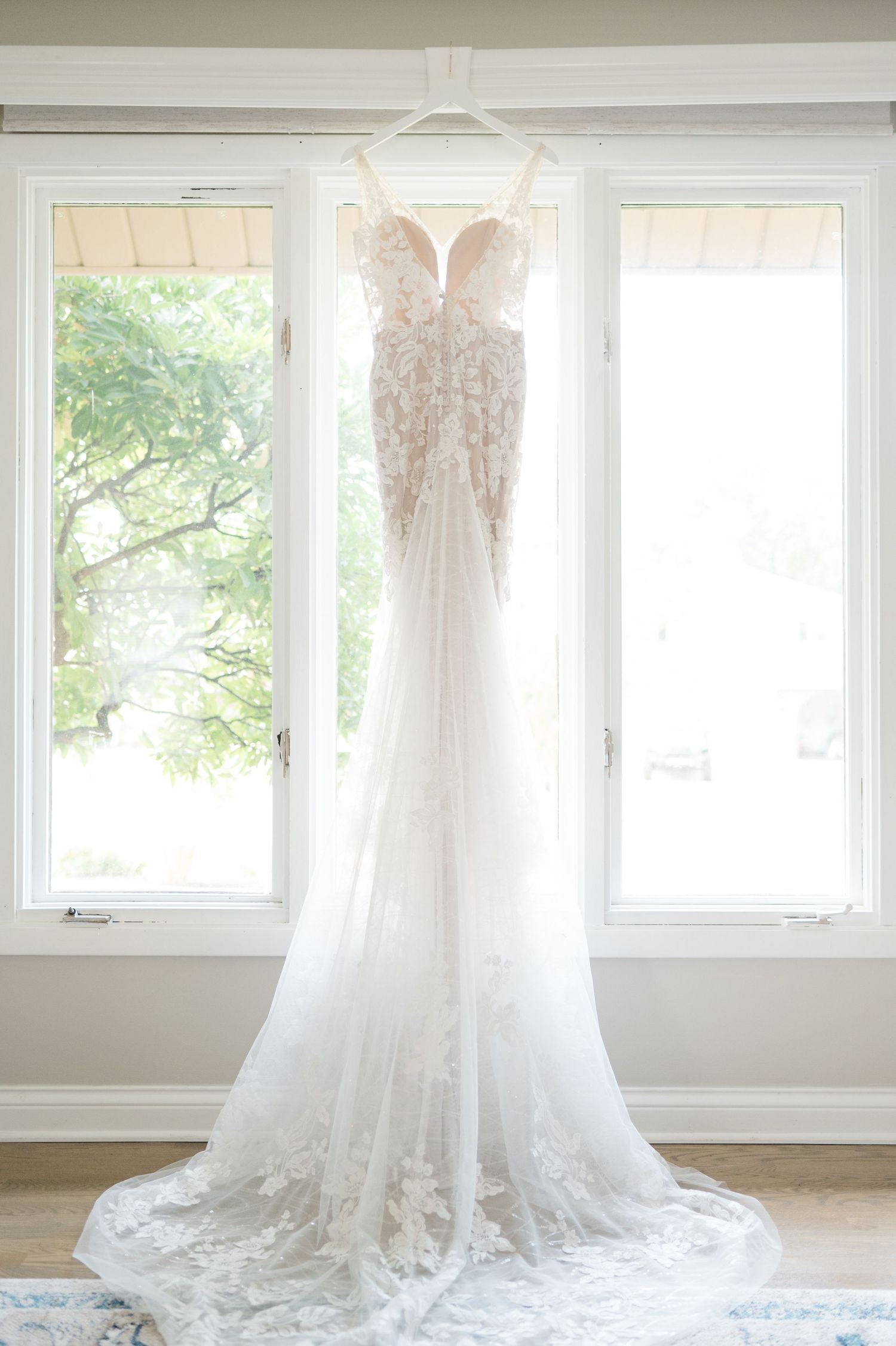 A delicate white lace wedding gown with sweetheart neckline hangs elegantly against bright windows in natural light.
