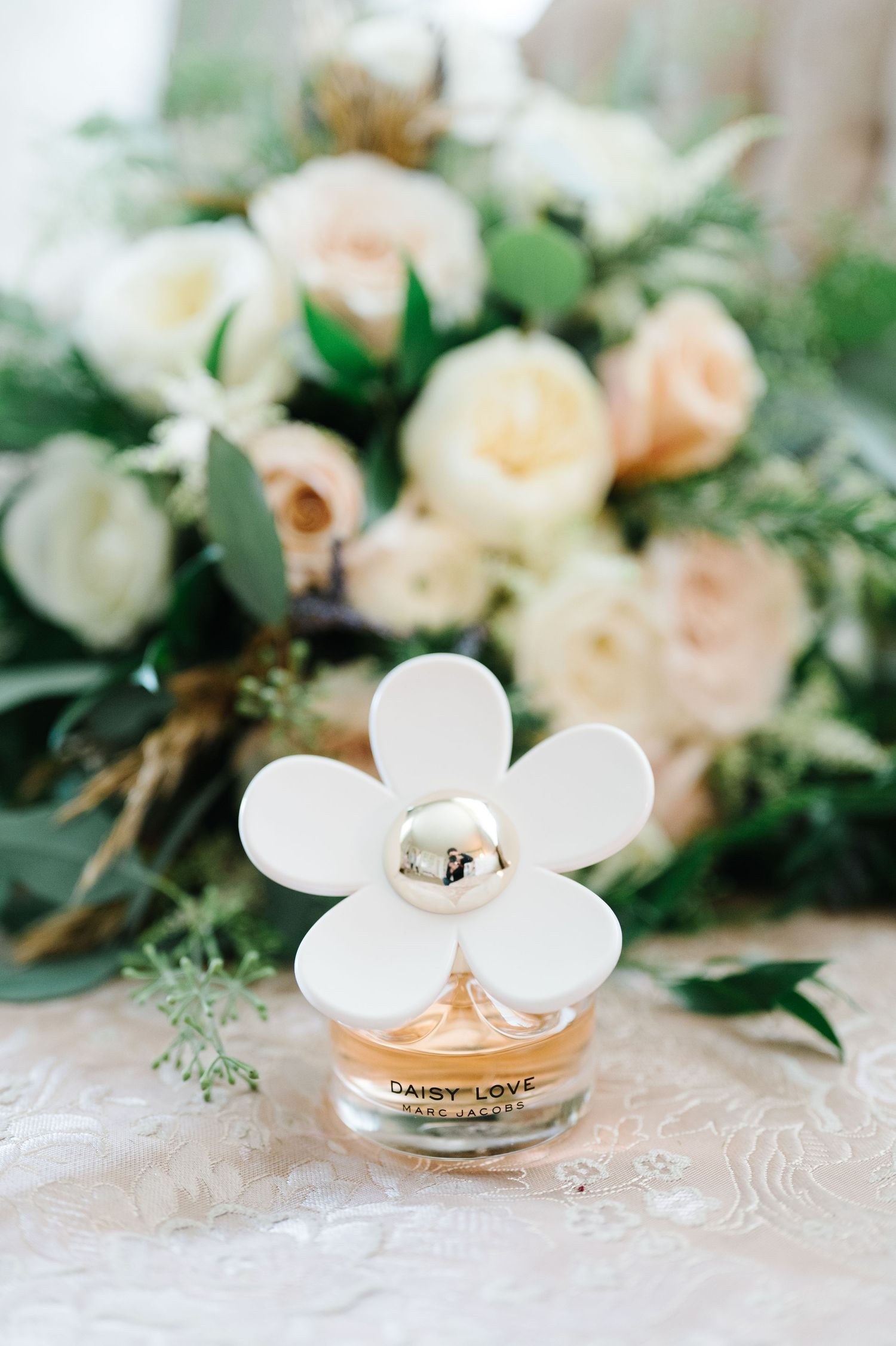 Wedding perfume bottle decorated with white flower petals sits among soft peach and cream roses and greenery.