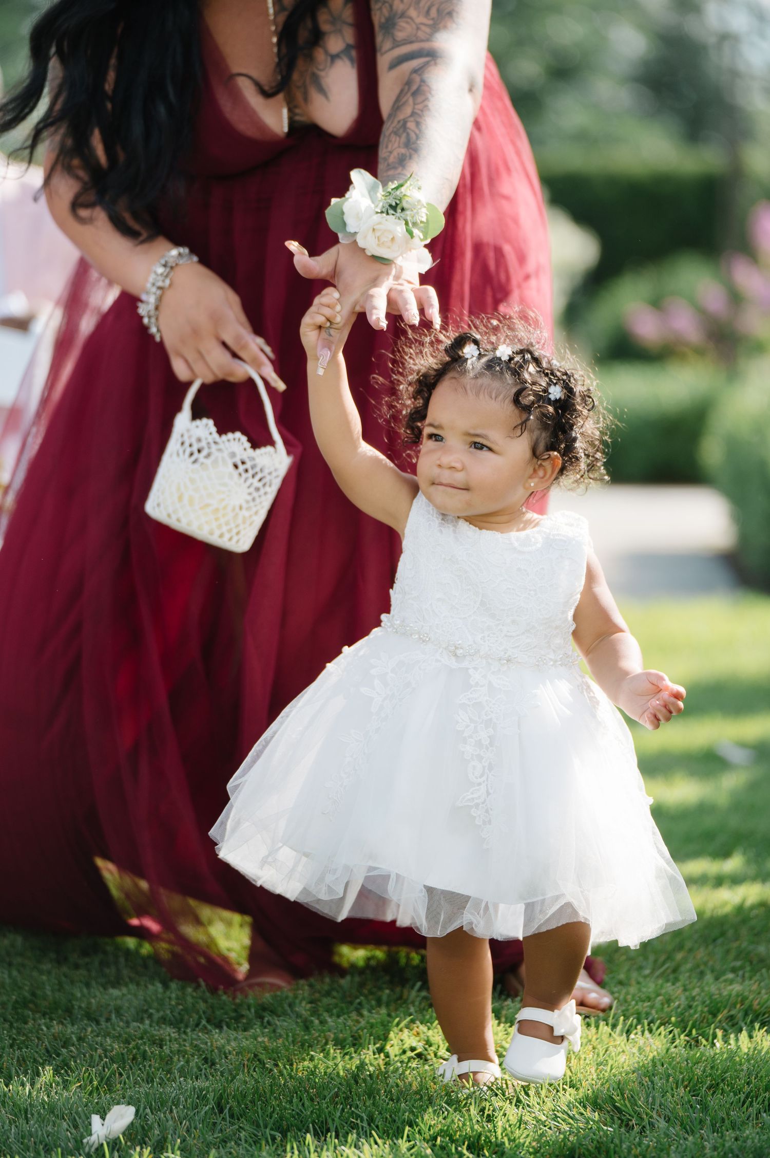 A young child in a white tulle dress holds hands with someone in burgundy at an outdoor wedding celebration.