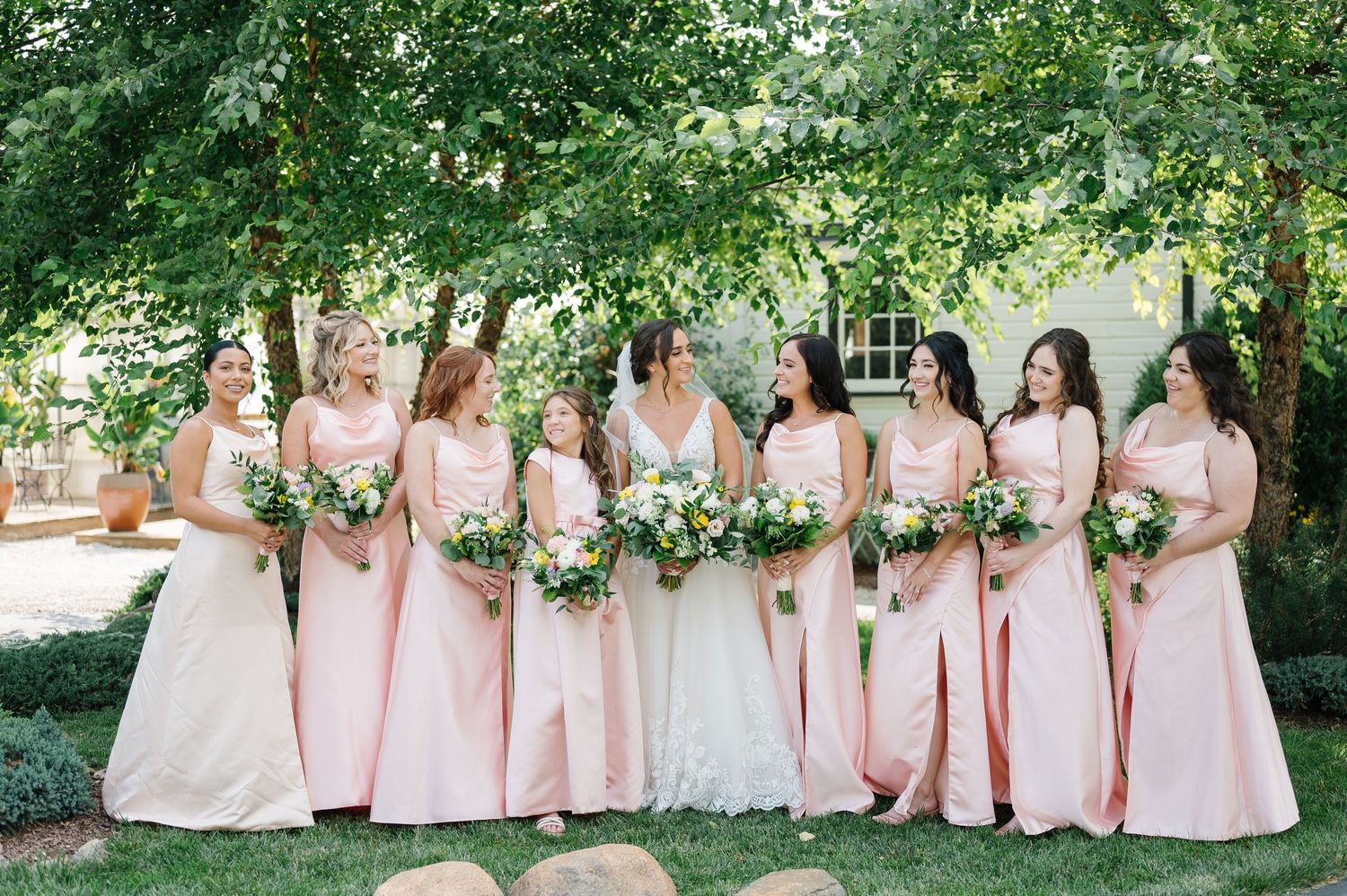 A wedding party in blush pink gowns stands in a line outdoors holding floral bouquets under green trees.